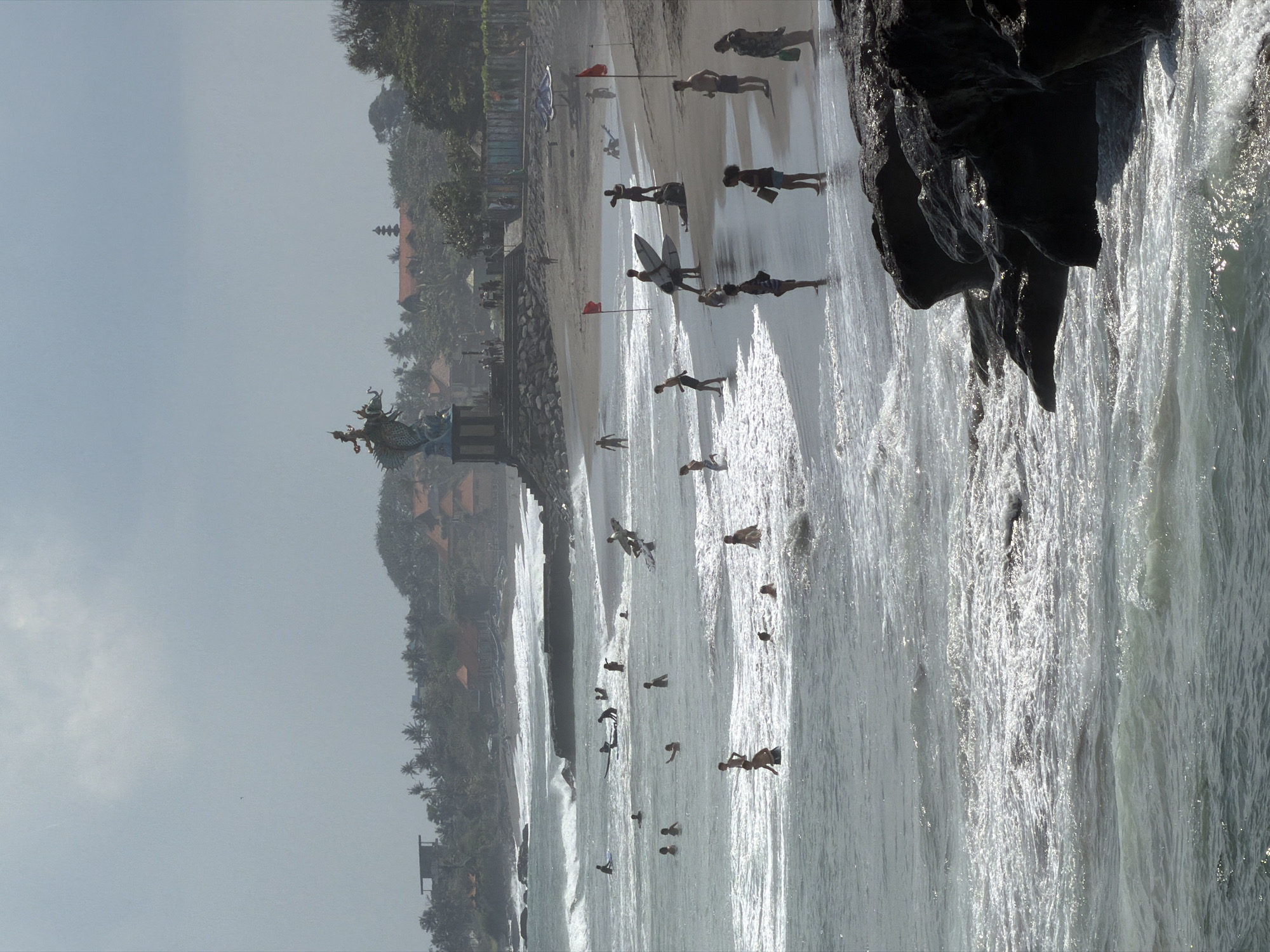 This is a medium shot, vertical, eye-level, outdoor photograph of a beach in North Kuta, Indonesia. The scene is dominated by a hazy sky and ocean. In the foreground, dark, wet rocks are visible on the right, with waves crashing against them. The ocean is choppy with white foam from breaking waves, and several people are wading or swimming in the water. Some are standing, others appear to be moving through the waves.
The beach itself is visible beyond the water, with a scattering of people. Some are standing on the sand, others are walking. Several individuals are holding surfboards, and at least one person is sitting on the sand. Red flags are planted in the sand at intervals.
In the middle ground, a large, ornate statue of a dragon-like creature with a rider on its back stands atop a stone pedestal. Behind the statue, lush green foliage and red-roofed buildings are visible, suggesting a developed area behind the beach. The overall atmosphere is somewhat subdued due to the overcast sky and haze, but the presence of people suggests an active recreational area.