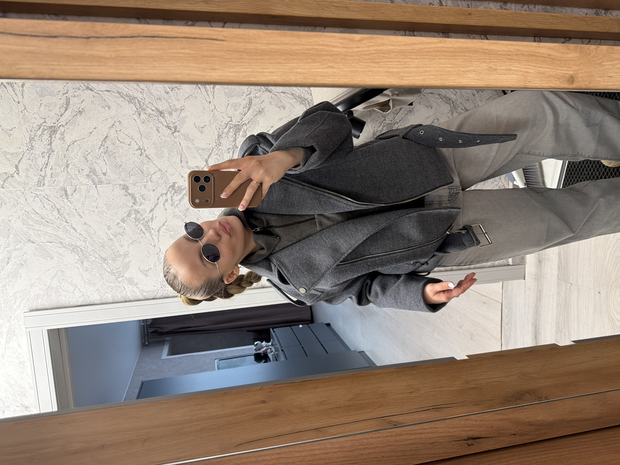 An indoor mirror selfie depicts a young female in Reshetnikovo, Russia. She is shown from the mid-thigh up, posing with her head slightly tilted and lips pursed. She wears dark round sunglasses, a dark grey asymmetrical zippered jacket, a grey-olive high-collared zipped top, and grey loose-fitting trousers. Her hair is pulled back in a ponytail with braids visible at the sides, and she has light-colored nail polish and a ring on her left ring finger. She holds a brown-cased smartphone with a triple-lens camera system in her right hand, positioned to capture her reflection. The background visible through the mirror shows a room with light-colored, patterned wallpaper. Reflected behind her, another room is visible through a doorway, featuring a dark chest of drawers with a mirror and decorative items, and dark curtains. The flooring appears to be light wood laminate or tile. A wooden door frame is prominent on the left and right sides of the reflection, and a white broom is visible on the floor near a dark mat in the bottom right corner of the reflected scene.