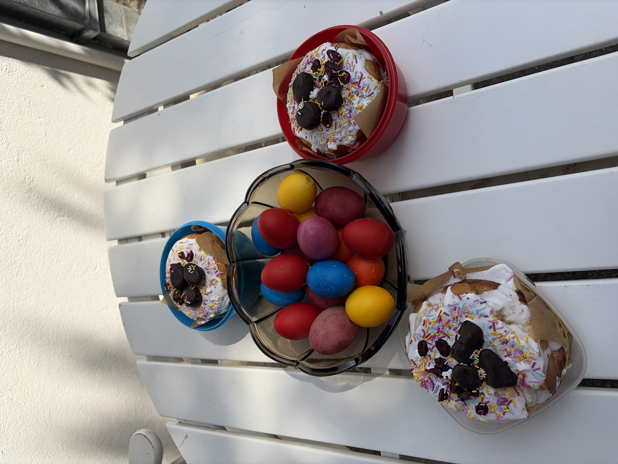 The image shows a white, slatted table with Easter treats arranged on it. There are three round cakes decorated with white icing, sprinkles, and dark chocolate pieces. One cake is in a red container, one in a blue container, and one in a clear square container. In the center of the table, a dark, bowl-shaped container holds a collection of colorful Easter eggs in shades of red, blue, yellow, purple, and orange. The setting appears to be outdoors, possibly on a patio. The location is in Topola Municipality, Serbia.
