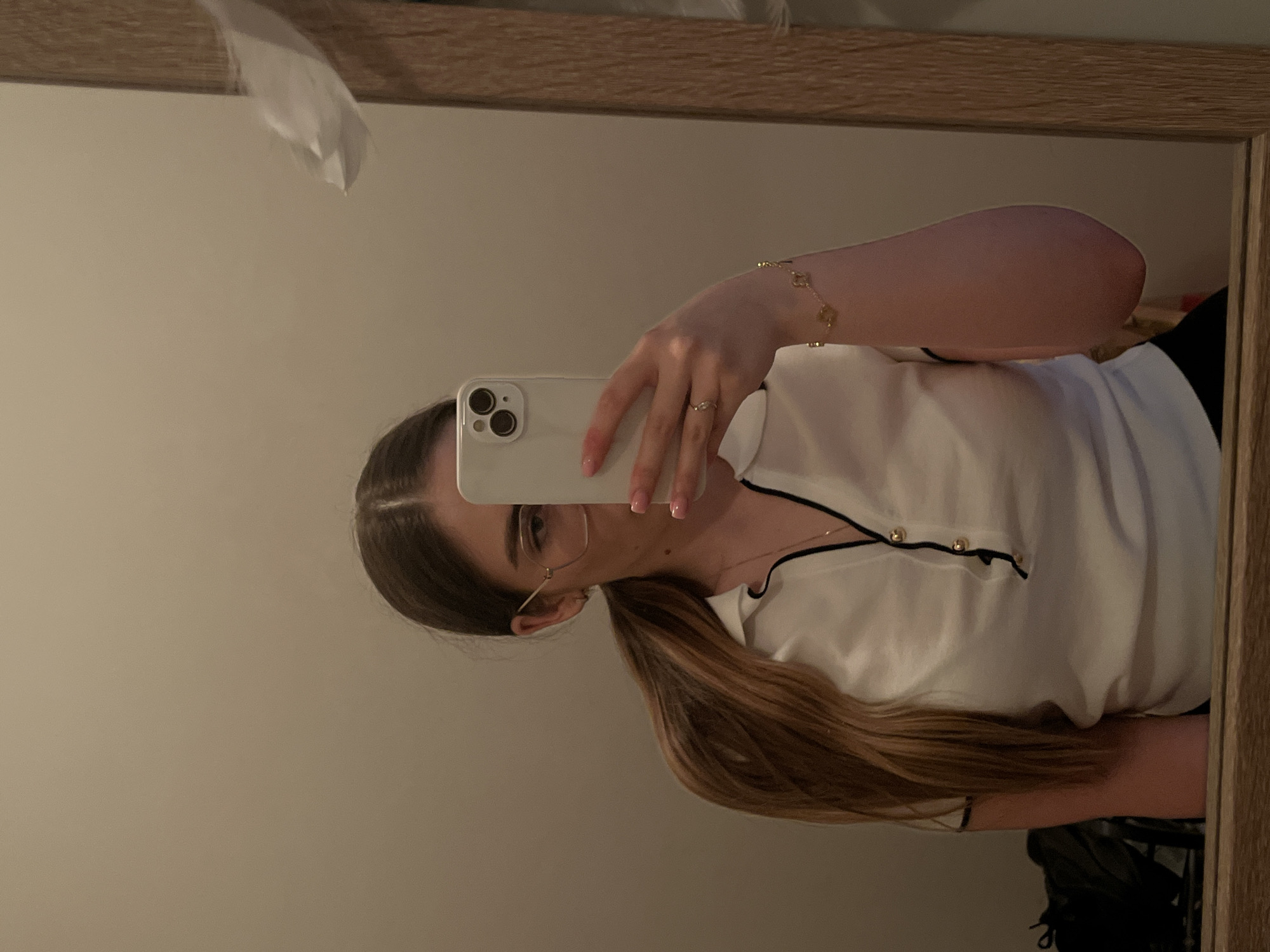 A mirror selfie captures a young adult female in an indoor location within the city of Unknown, Unknown. The subject has long, straight, brown hair tied in a low ponytail visible on her left side. She wears gold-rimmed rectangular glasses, and her face is partially obscured by a white smartphone held in her left hand. Her left hand's ring finger displays a thin gold-colored ring, and her fingernails have light pink polish. She is dressed in a white short-sleeved collared shirt with black trim on the collar and placket, fastened with two visible gold-colored buttons. A delicate gold-colored necklace with a small pendant is visible around her neck, and a gold-colored bracelet with four clover-shaped charms adorns her right wrist. Dark-colored bottoms are partially visible at the bottom of the frame. The background, reflected in the mirror, is a plain, light brown wall. A wooden mirror frame runs vertically along the right side. In the lower-left of the reflection, a dark object, possibly a bag, is visible. In the upper-right, a crumpled white fabric item is reflected near the mirror frame.