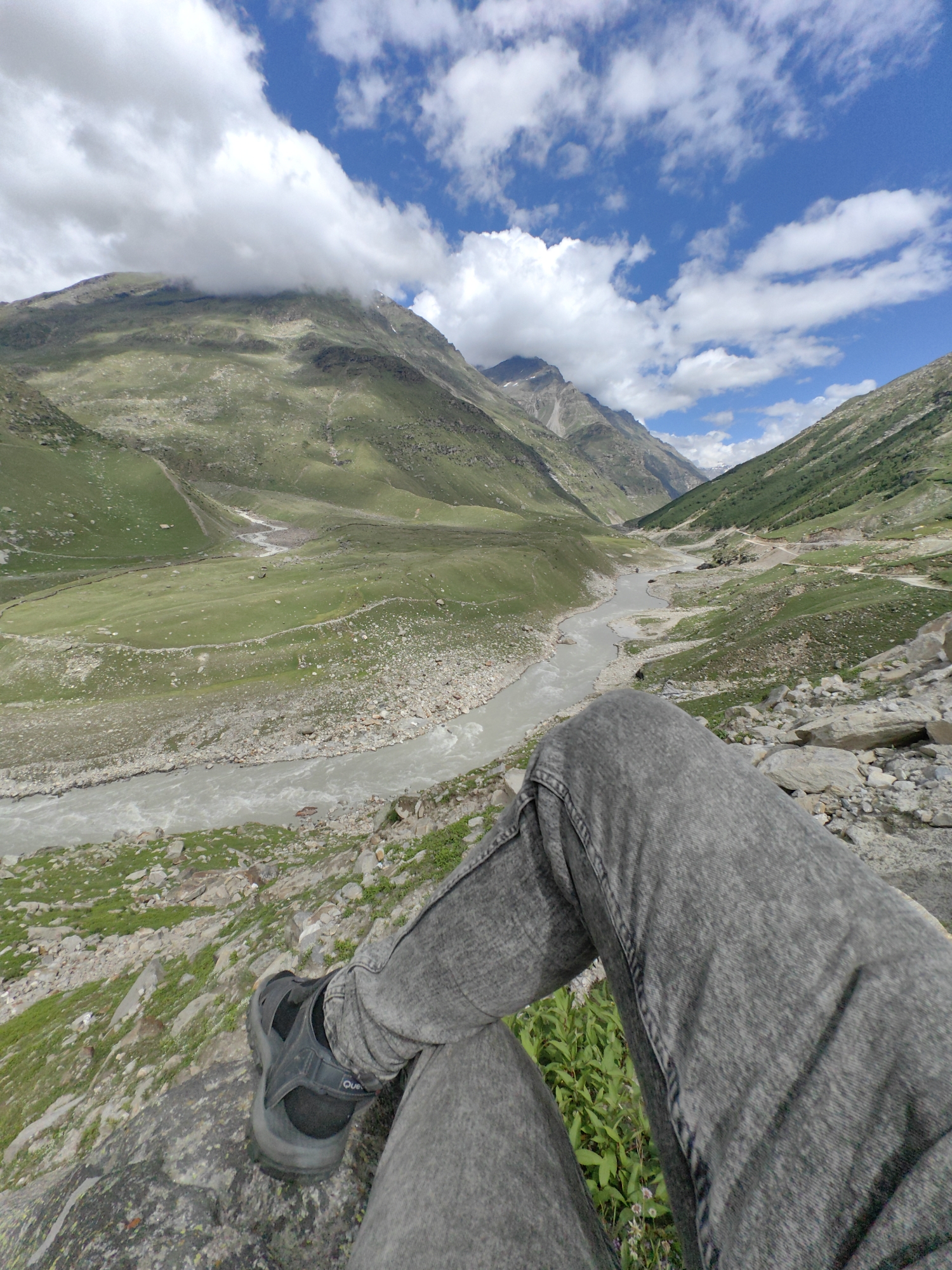 The image shows a scenic view of a valley in Burwa, India, featuring a river flowing through a green landscape with rolling hills and mountains under a partly cloudy sky. In the foreground, a person's legs, clad in grey jeans and a black sandal, are visible as they sit on a rocky outcrop overlooking the valley. The river appears to be wide and somewhat turbulent, with a rocky bank on the right side. The mountains in the background are covered in green vegetation, with some areas appearing rocky and barren. The sky is a vibrant blue with scattered white clouds, some of which are obscuring the peaks of the distant mountains. The overall impression is one of natural beauty and tranquility.