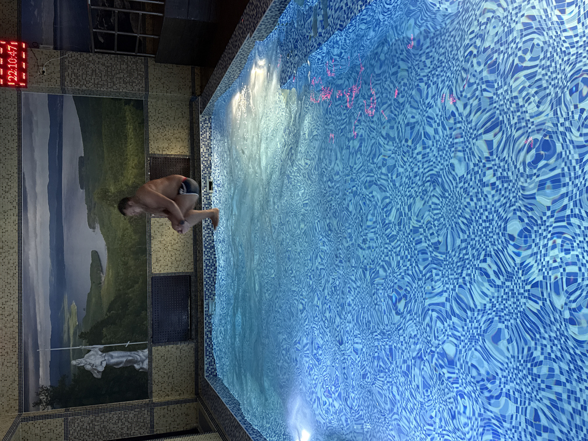 A man is diving into a blue-tiled indoor pool, potentially located in Zolotoi Plyazh, Russia. The pool is surrounded by mosaic tiles, including a mural of a landscape scene and a statue. A digital clock is visible on the wall.