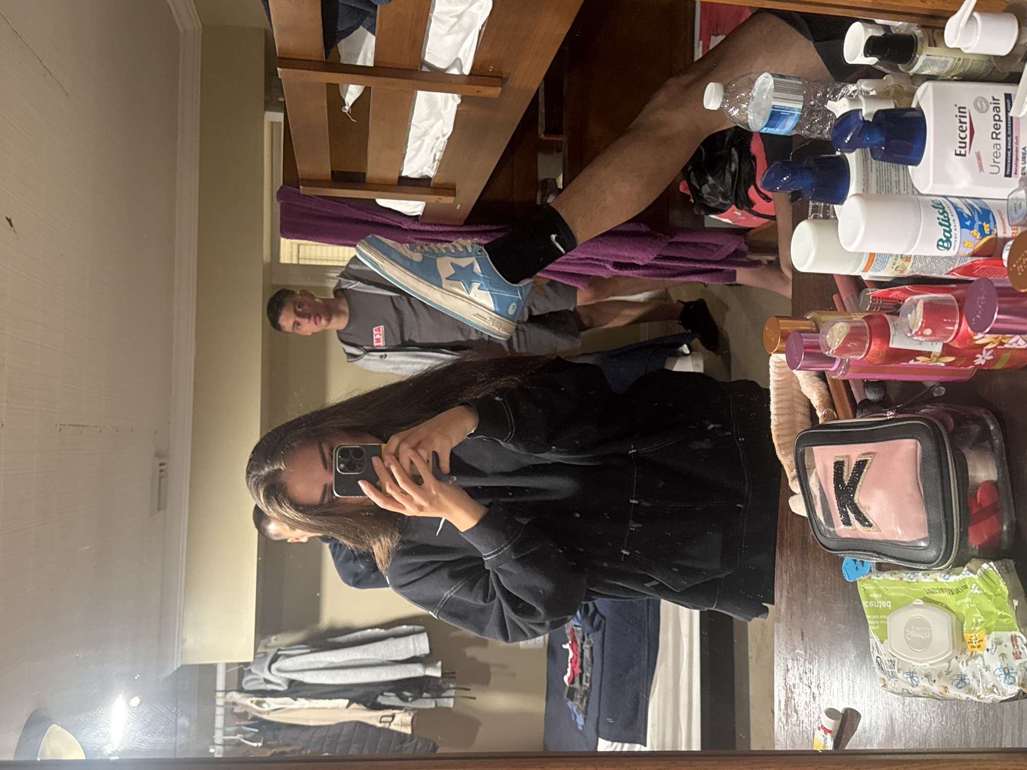 This is a mirror selfie taken in a dorm room, likely at a motel or hotel in Ocean City, Maryland. In the foreground, a young woman is taking the photo with her phone, wearing a black hoodie. Her long brown hair obscures part of her face. To her right, in the reflection, a young man in a grey t-shirt and shorts stands near a bunk bed. Another young man is partially visible behind the woman. A third person's legs are visible in the reflection, with one leg raised and wearing a blue and white sneaker. On the table in front of the mirror are various toiletries, including baby wipes, a makeup bag, and several bottles of lotion and hair spray. The room appears to be a shared living space with bunk beds and a closet filled with clothes.