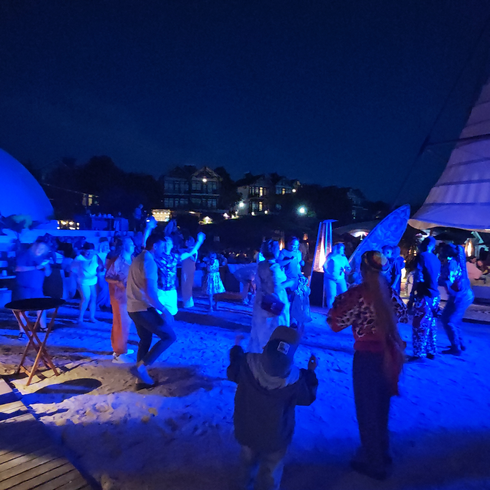 An outdoor nighttime party is in progress on a sandy area, possibly a beach, with blue lights illuminating the scene. In the background, a cluster of multi-story buildings with illuminated windows is visible on a dark hillside, indicative of a settlement within the city of Shchyolkino, Ukraine. Several people are scattered across the sand, some dancing. In the foreground, a person wearing a dark jacket and hat is facing away from the camera, appearing to dance or move. To the left, a small, round table with a tripod base sits on the sand. A large, white tent-like structure is partially visible on the right side of the frame.