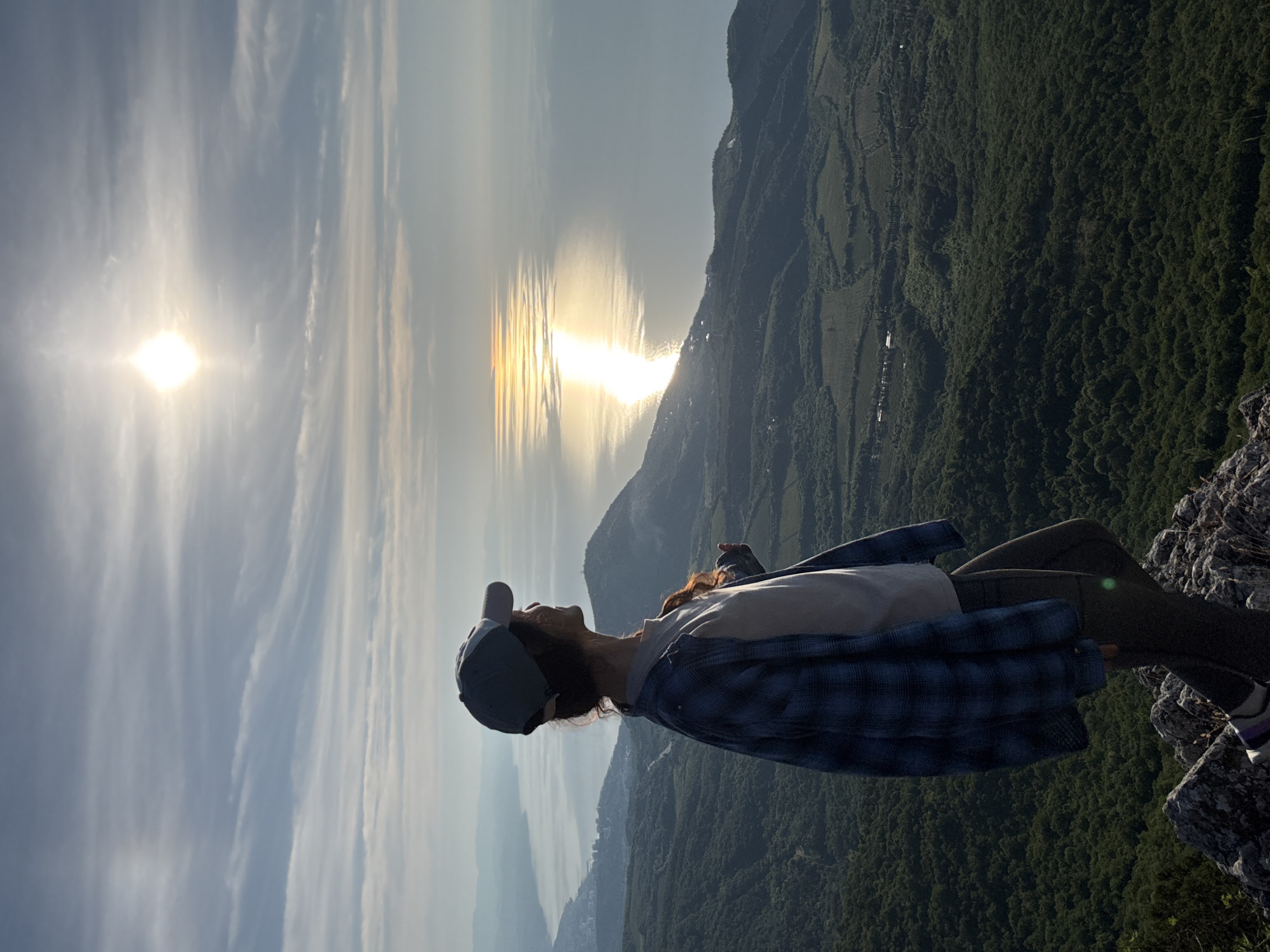 A lone individual stands on a rocky mountain peak overlooking an expansive landscape. The person, viewed from the back and right side, wears a grey baseball cap, a light-colored t-shirt, a blue and black plaid long-sleeved shirt worn open, and dark grey or black pants or leggings. Striped socks (purple, white, grey) are visible on the left foot. Reddish-brown hair is visible beneath the cap, catching sunlight. The individual's head is turned to the right, looking out over the scene.
The foreground consists of grey rocks and sparse vegetation. Beyond, dense dark green forests cover rolling hills and valleys. Patches of lighter green fields, possibly agricultural, are interspersed within the forested areas. In the mid-distance to the left, a coastal city or town with numerous buildings extends along the shoreline. A large body of water, likely a sea or ocean, stretches across the horizon. The sun is low in the upper right sky, casting a brilliant, elongated reflection path across the water, extending towards the lower right. The sky is partly cloudy with thin, wispy cloud formations. The lighting indicates either sunrise or sunset. The specific location is Unknown, Unknown.