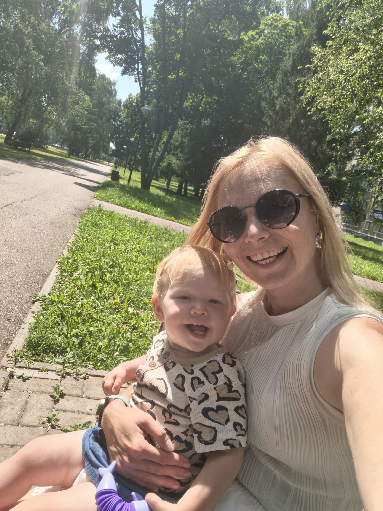 A blonde woman wearing a white sleeveless top and round sunglasses smiles at the camera, holding a young child in her lap. The child, with blonde hair and wearing a leopard-print shirt, is also smiling. They are sitting on a bench outdoors, with a paved path and lush green grass visible in the background, bordered by trees. Sunlight streams through the trees, creating dappled light. The scene appears to be a park in the city of Sterlitamak, Russia, likely during the daytime.