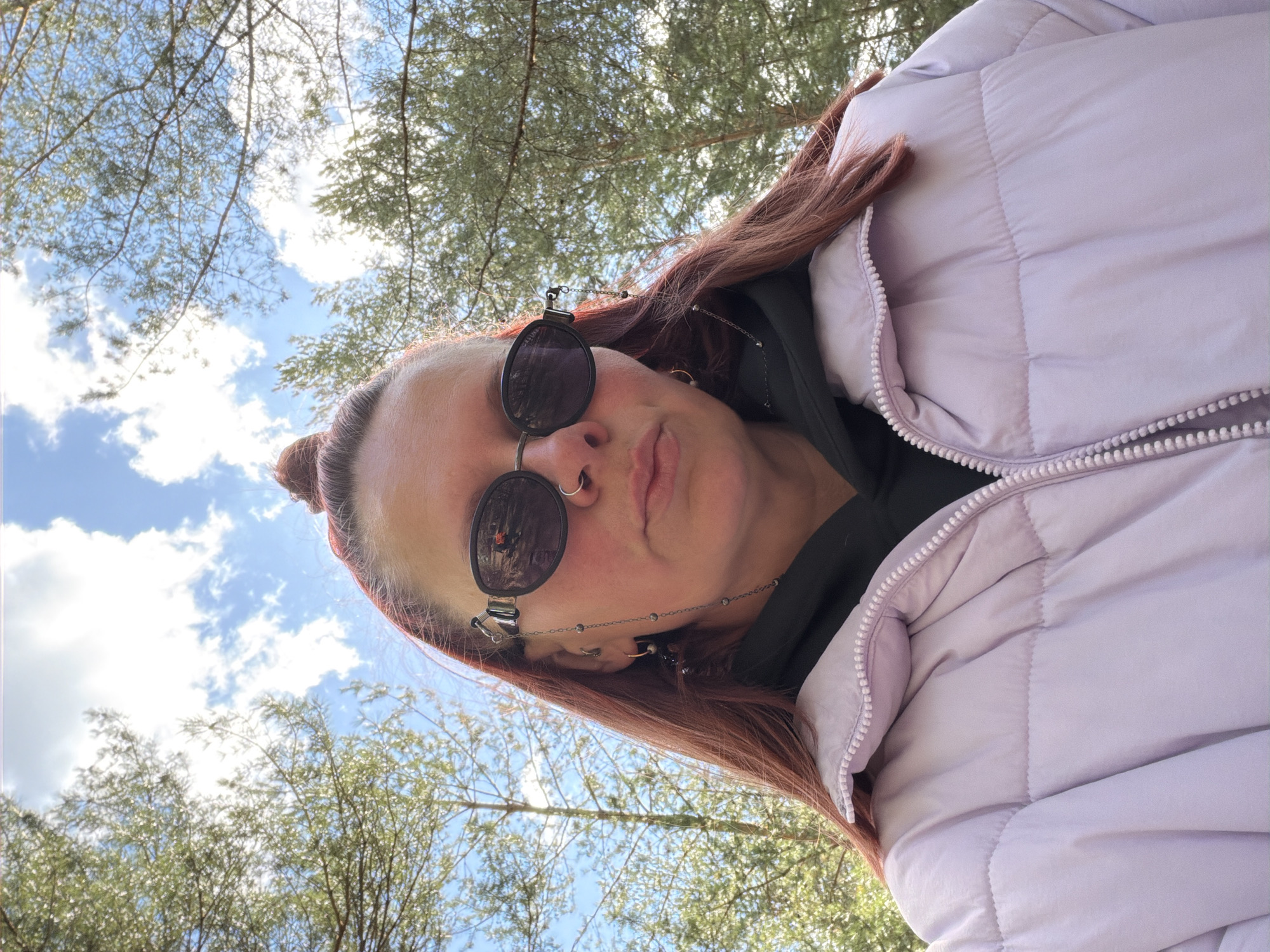 A woman with reddish-brown hair in a bun is wearing a purple puffer jacket, a dark hoodie, and round sunglasses with a beaded chain. She is looking at the camera from a low angle, with trees and a blue sky with white clouds visible in the background. The location appears to be outdoors. Location in Chaikovsky, Russia cannot be confirmed.