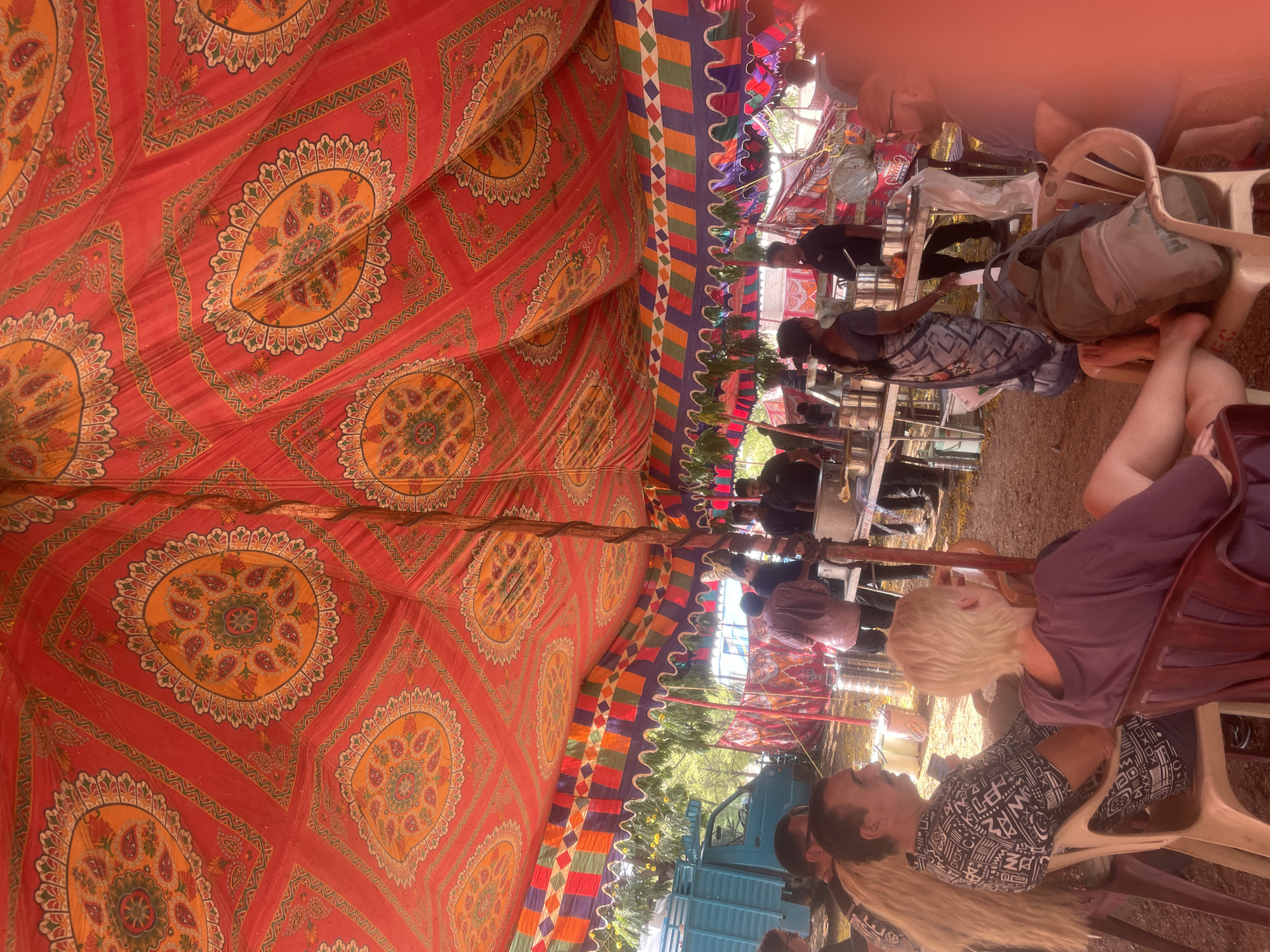 The image shows a partially visible outdoor scene in Anajpur, India, under a large, ornate red tent with geometric and floral patterns. The tent's edges are decorated with colorful stripes. People are seated on plastic chairs, with some individuals visible in profile, wearing casual clothing.
A light blue truck is visible outside the tent's perimeter. Tables are set up within the tent, and some individuals appear to be serving food or beverages. The overall atmosphere suggests a gathering or event. The ground is partially visible and appears to be an outdoor area.