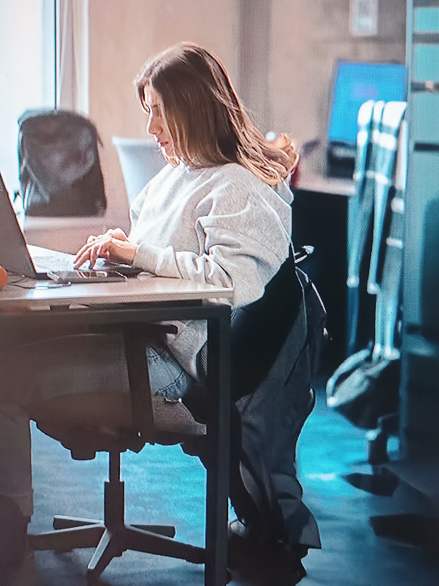 A woman with shoulder-length brown hair sits at a desk in an office setting, typing on a laptop. She wears a light grey sweatshirt and blue jeans. A black backpack is behind her on the chair. A second backpack is visible behind her and to the left. A computer monitor can be seen in the background and to the right. The office has blue lighting. The location is not identifiable as Tbilisi, Georgia based on the visual information provided.