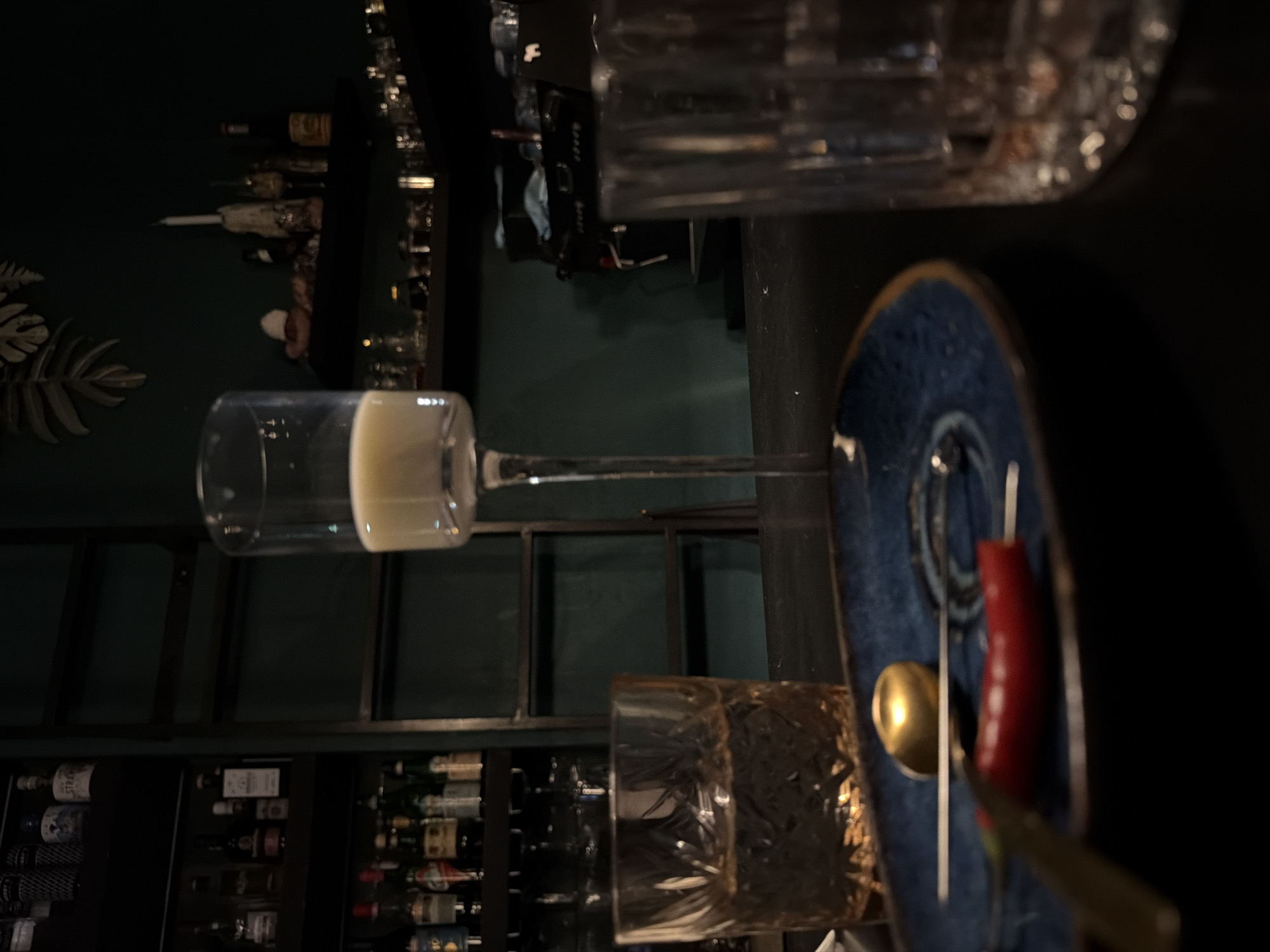 The image shows an indoor bar setting. In the foreground, there's a blue decorative plate with a gold spoon and a red chili pepper. Two glasses are also visible: one with a clear liquid and another with a pattern on its surface. The background features a bar shelf stocked with various bottles, likely alcohol. The color palette leans towards dark greens and browns, creating a dim and intimate ambiance. No people are discernible in the frame. The setting appears to be a sophisticated cocktail bar, but the location is Unknown, Unknown.