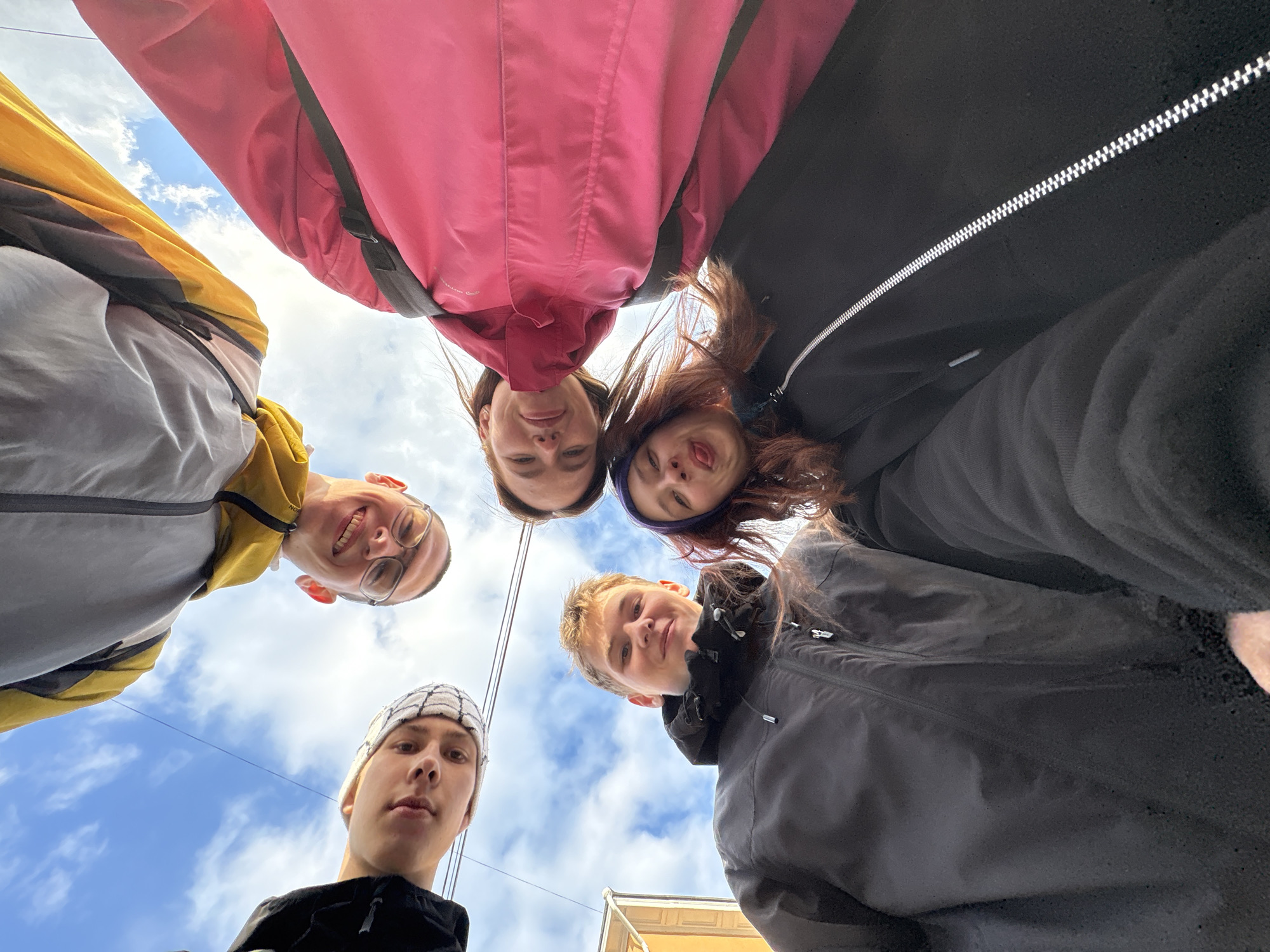 Five young people are in the city of Perm, Russia, standing close together in a circle, looking down at the camera. The angle is from below, pointing upwards at their faces. The sky with scattered clouds is visible in the background. The individuals are wearing casual outerwear, including jackets and a beanie. A building facade with a yellowish hue is barely visible at the bottom right corner. The lighting is natural, suggesting it is daytime. One individual is sticking out their tongue.