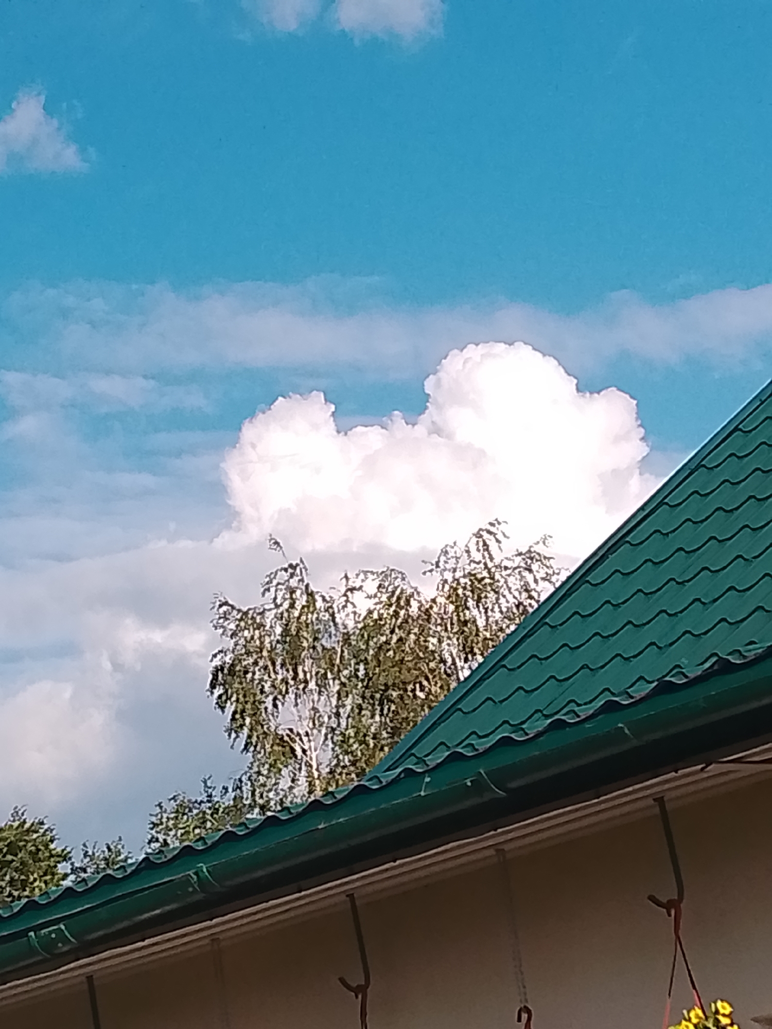 The image shows a cloudy sky with a large, bright cloud formation resembling a heart shape. In the foreground, a green, wavy metal roof of a building is visible, along with the edge of a light-colored wall. Behind the roof, the tops of trees with green foliage are seen against the sky. The scene appears to be in an outdoor setting, possibly in a rural or suburban area within the Свіслацкі сельскі Савет, Belarus, given the typical architecture and natural elements. There are no people or specific actions depicted in the image.