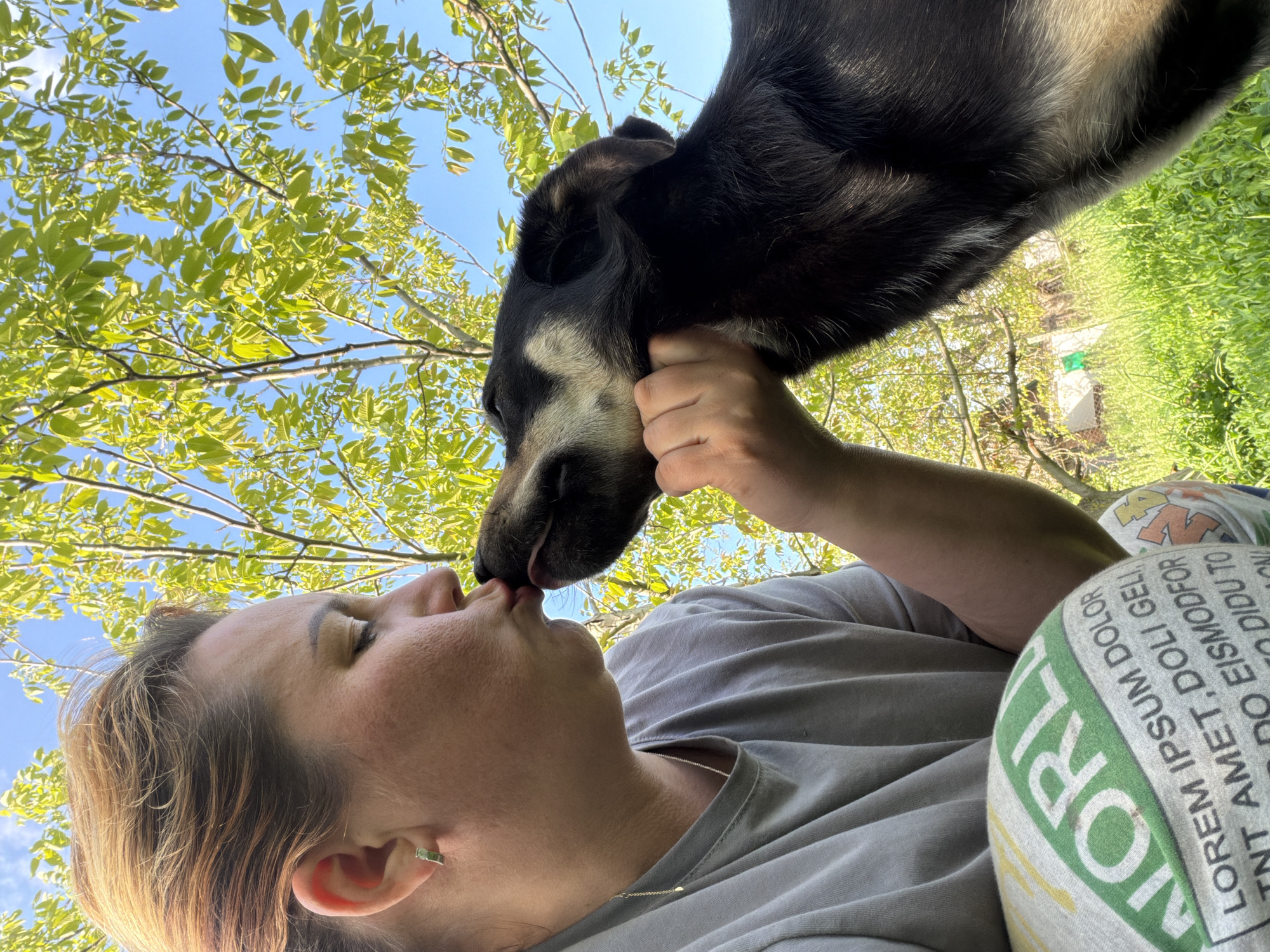 A low-angle shot shows a woman receiving a kiss from a dog, possibly outdoors in Leningradskaya, Russia. The woman, wearing a gray t-shirt and printed pajama pants, closes her eyes and accepts the dog's affection. The dog is black and tan, its tongue extended towards her face. Lush green foliage and a clear blue sky with tree branches overhead form the background. The lighting suggests it is daytime. The woman is holding the dog's face with one hand.