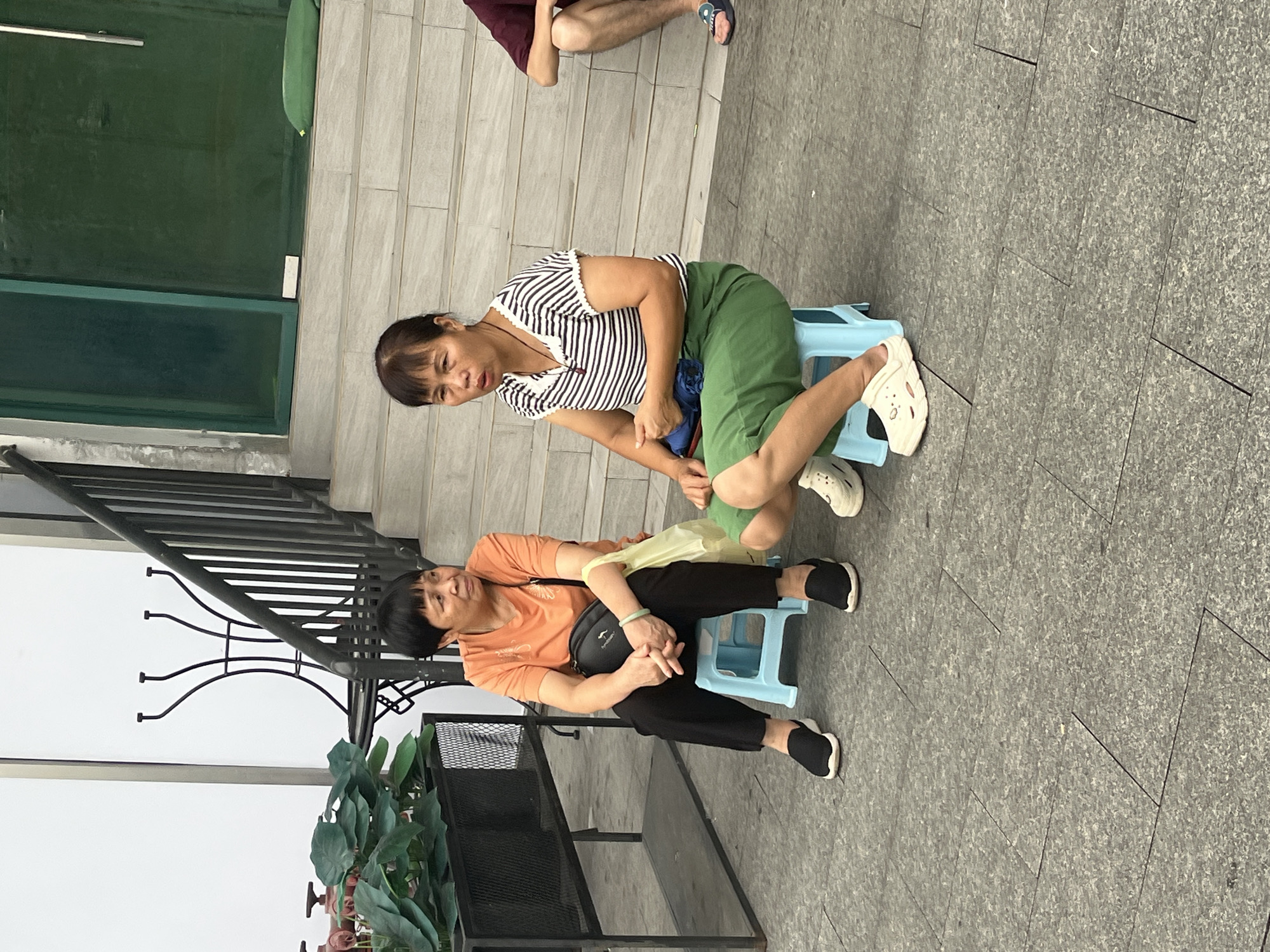 Two adult Asian women are sitting on light blue plastic stools on a grey tiled outdoor area. The woman on the left is wearing an orange short-sleeved t-shirt and black pants, with black slip-on shoes. She has a black shoulder bag and is holding a light yellow plastic bag in her lap. The woman on the right is wearing a black and white striped short-sleeved shirt and a green skirt. She is wearing white clogs. She has a blue bag in her lap. In the background, there is a staircase leading up to a building with a green door. To the left of the staircase is a black metal planter with green foliage. The scene appears to be in the Chancheng District of China.
