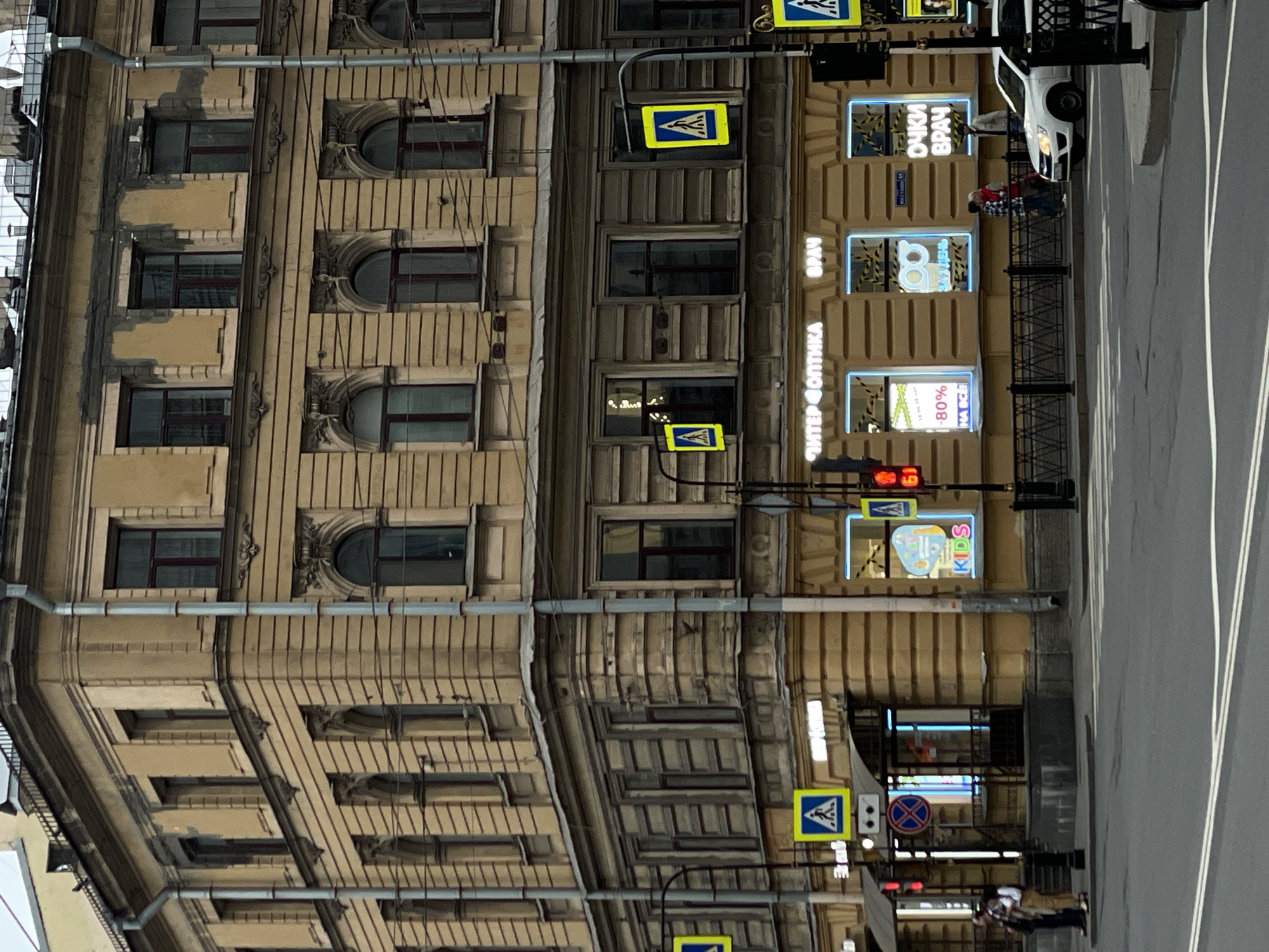 Here is a detailed description of the image:
**Scene Overview:** The image is a street scene in what appears to be Saint Petersburg, Russia, judging by architectural style and signage. The perspective is slightly tilted and appears to be taken from a position above street level.
**Architecture and Buildings:** The focus is on a tall, ornately decorated building with multiple windows. The building is likely of historic significance and has a facade with details such as molded frames around the windows and vertically fluted sections. There's another building visible, a shorter structure next door with advertising signage.
**Signage and Advertising:** The taller building features a vertical sign that reads "ПИТЕР ФОПТИКА" with additional advertising for an "ОЧКИ ВРАЧ" (Eye Doctor) and various promotions like "-80% HA BCE" (80% Off Everything). Another sign reads "KIDS" and "ЗА 1 ДЕНЬ" (In 1 Day).
**Street and Road:** The right side of the image shows a street with visible lane markings. There are a few vehicles on the street, including what appears to be a white car.
**People and Activities:** There are a few people visible on the sidewalk, including what looks like a couple in front of the advertisement window. Further in the distance, there is a person walking on the side walk.
**Other Objects:** The scene includes pedestrian crossing signs with the characteristic blue triangle and pedestrian icon, traffic lights. The street is bordered by a metal fence.