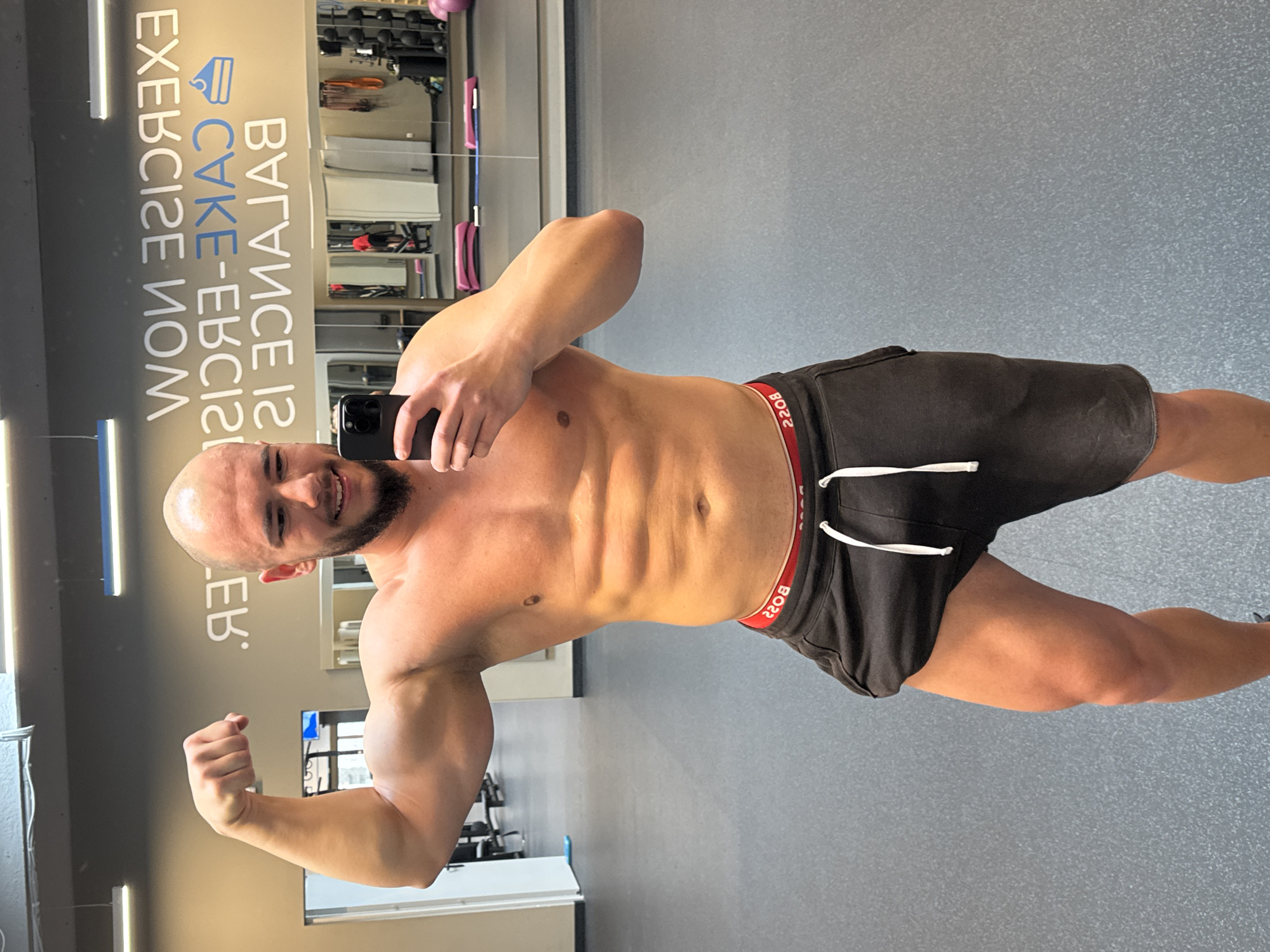 A man is taking a selfie in a gym. He is shirtless and wearing black shorts with a red waistband. He is flexing his left bicep and smiling. The gym has gray walls and floors. There is a mirror in the background with text that reads "BALANCE IS CAKE-ERCISE NOW." There are gym equipment visible in the background. The location is a gym and based on the gym equipment, is most likely in a Fitness center in any city, not specifically Sofia, Bulgaria.