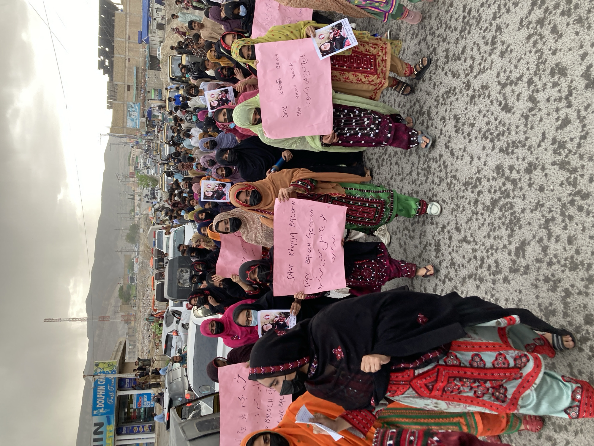 The image depicts a large outdoor protest or demonstration taking place on a paved road, likely in Quetta City Tehsil, Pakistan, under an overcast sky during daylight. The primary subjects are a numerous group of people, predominantly women, extending into the background.
Many of the women are dressed in traditional Balochi attire, featuring colorful, embroidered dresses, and most wear head coverings; some also wear face masks. They are actively engaged in the protest, holding up pink placards with messages written in both English and Urdu. Visible English text on the placards includes "SAVE KADIGA BALOCH", "SAVE Khaija BALOCH", and "Stop BALOCH Genocide." The corresponding Urdu text, "بلوچ نسل کشی بند کریں" (Baloch nasal kushi band karen), also translates to "Stop Baloch Genocide." Some individuals are holding small printed photos, with text like "Save Khadija Baloch" and "Missing from Balochistan."
In the background, a street scene unfolds with various vehicles, including cars and rickshaws, and multi-story buildings. Distant mountains frame the horizon. A shop sign on the left side of the image reads "DOLPHIN CH Fast Food" and "Hico DAIRY ICE CREAM." The gathering conveys a sense of collective concern and demand for action regarding the issues of Baloch genocide and the safety of individuals like Khadija Baloch.