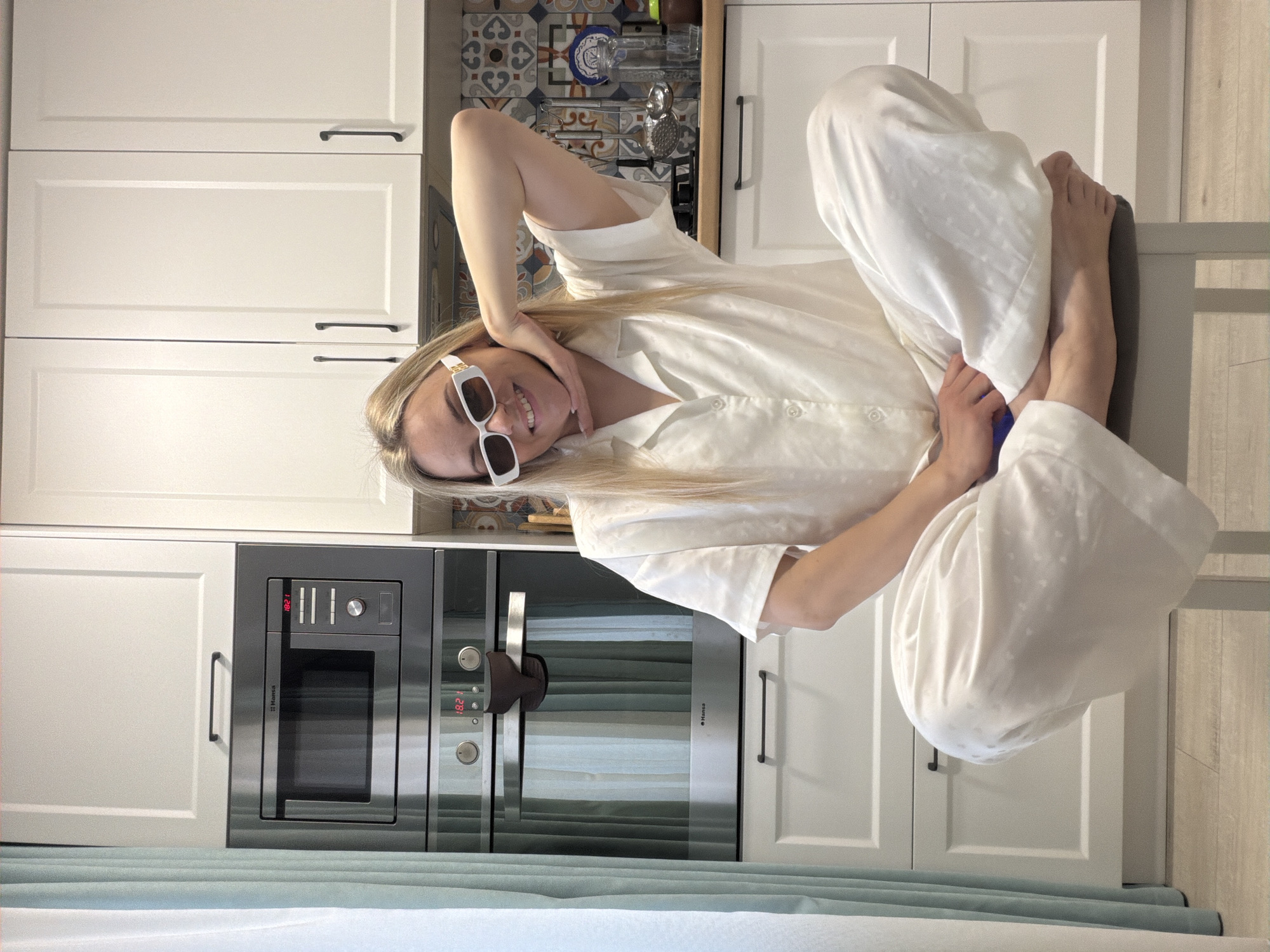 A young woman with long blonde hair and a bright smile poses playfully in a modern kitchen. She is wearing white rectangular sunglasses, a white long-sleeved collared shirt, and matching wide-leg pants, suggesting comfortable loungewear. Seated on a dark grey stool or chair, she leans back against white kitchen cabinets, with one arm resting on the top cabinet and her bare feet propped up. Her pose is relaxed and cheerful as she looks directly at the camera.
The kitchen features clean white cabinets with dark handles, typical of contemporary design. Built into the cabinetry on the left are two stainless steel appliances, likely an oven and a microwave, both displaying the time "18:21" digitally, and one bearing the "Hansa" brand name. Above the woman's head, an open upper cabinet reveals a distinctive tiled backsplash with a geometric pattern in blues, browns, and whites, alongside various kitchen items like glassware and a metal grater. The floor appears to be light-colored wood or laminate. A light blue fabric, possibly a curtain or blanket, is visible in the foreground. The scene is brightly lit, indicative of daytime. This photograph captures a relaxed moment in a home kitchen in Votkinsk, Russia.