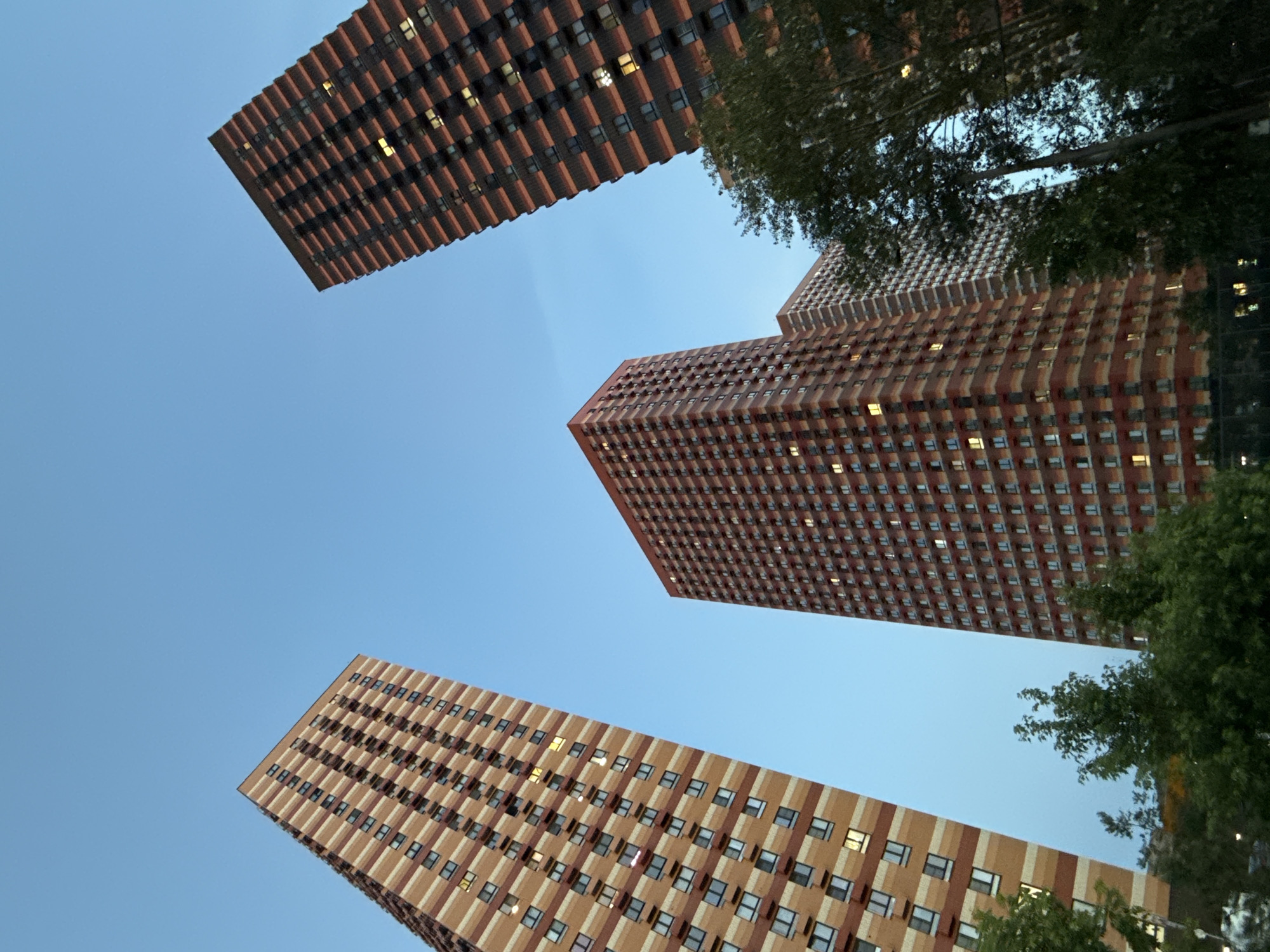 This image shows a low-angle view of several tall, modern apartment buildings against a clear, pale blue sky. The buildings have a distinctive facade with alternating vertical bands of reddish-brown and beige or light brown. Each building is densely packed with windows, and some of these windows are illuminated, suggesting it is either dusk or dawn, or that lights are on inside the apartments. Green trees with dense foliage are visible in the foreground and partially obscure some of the buildings, adding a natural element to the urban scene. The perspective is dramatic, looking directly upwards, which emphasizes the height and scale of the architecture. The overall impression is one of urban development and residential living. There is no visible text in the image.