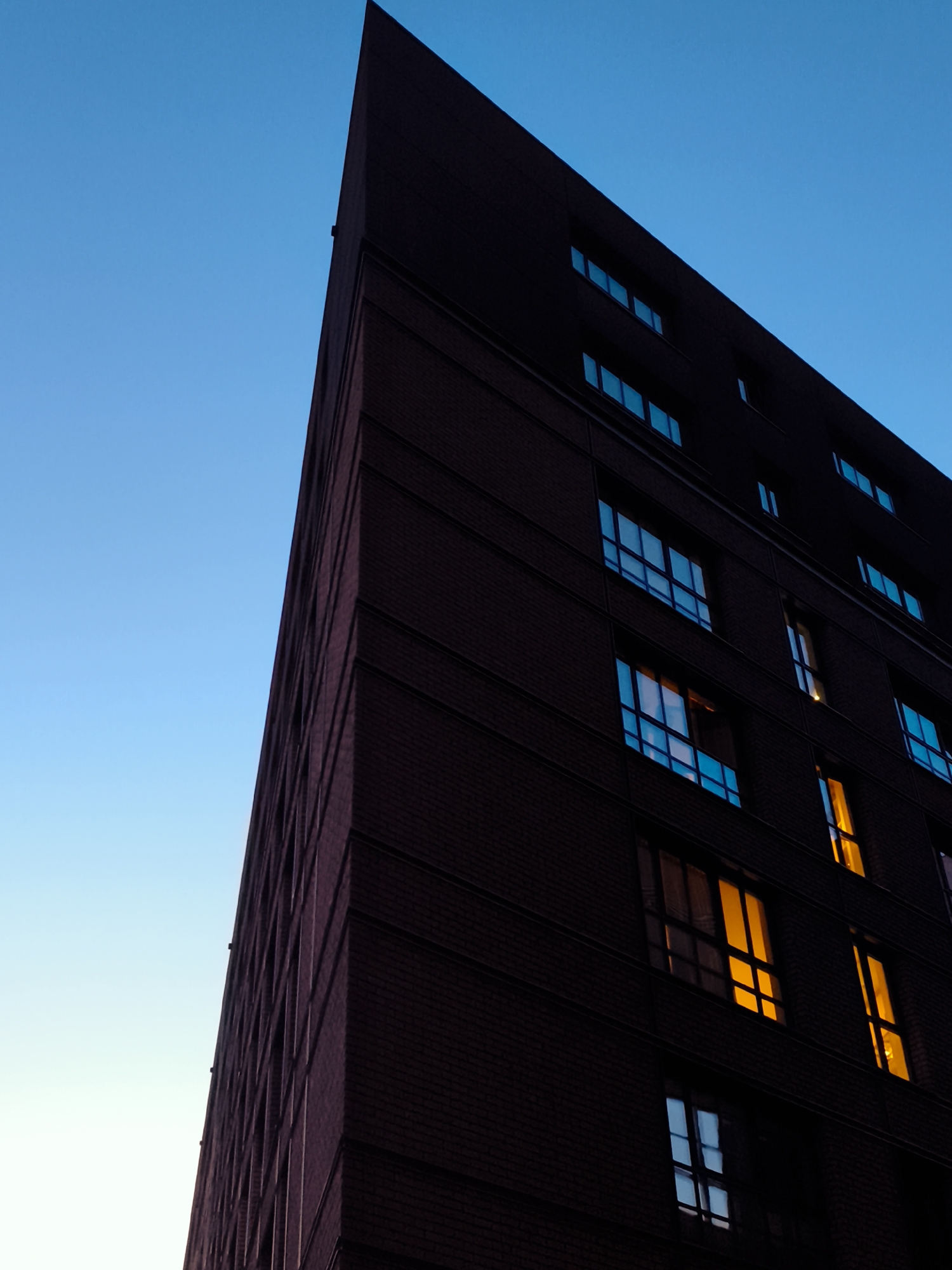 An upward, angled exterior shot shows the dark brick facade of a modern building against a clear, bright blue sky. The building has a sharp, triangular profile, with its apex pointing towards the top left of the frame. Numerous windows are visible on the building's surface. Some windows are dark and reflect the blue of the sky, while others emit a warm, yellow light, indicating interior illumination. The building is located in Gdańsk, Poland.
