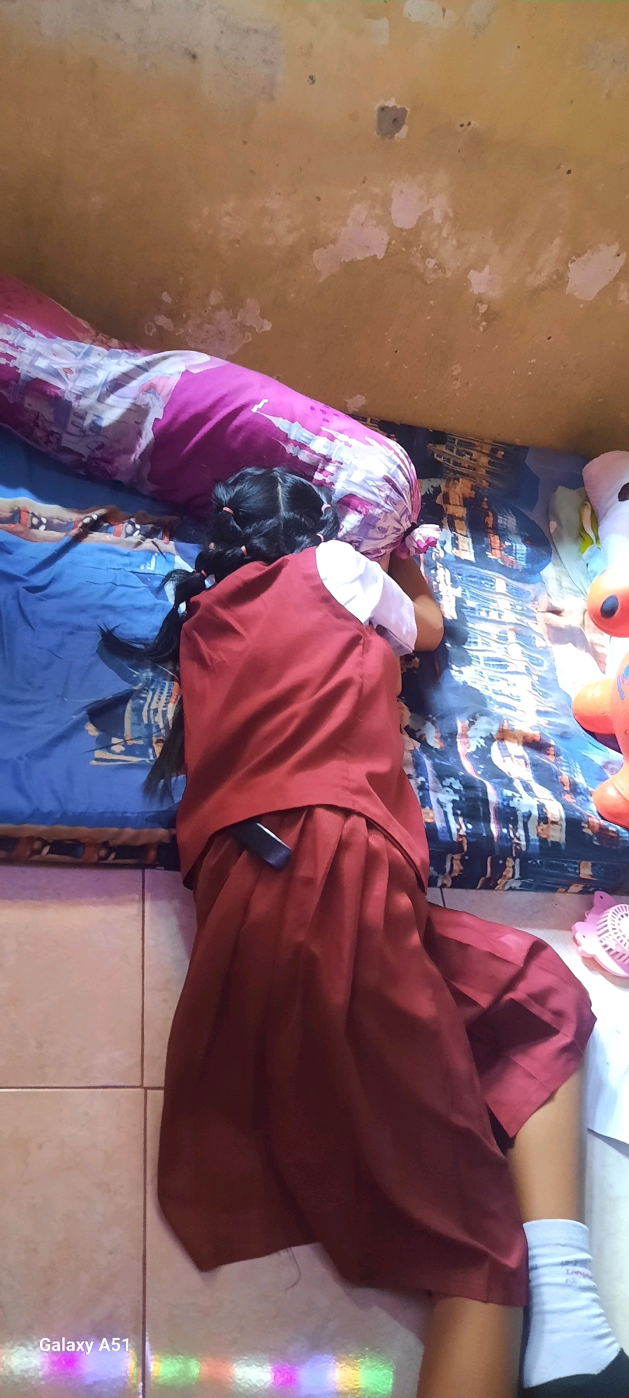 A person, likely a child, is lying on their stomach on a bed, facing a pillow with a castle design. The person is wearing a school uniform consisting of a maroon vest and skirt over a white shirt, with black hair styled in two braids tied with pink bands. A dark object, possibly a phone, is tucked into the waistband of the skirt. The bed is covered with a blue patterned sheet. In the background, a yellow wall shows signs of wear and peeling paint. To the right of the bed, there are plush toys, including a white one with black features and an orange one, as well as a pink mesh object. The floor is tiled with a beige-colored pattern, and a reflection of colorful lights is visible on the lower left portion of the floor. The image is tagged with "Galaxy A51". The context provided places this scene in Jatitengah, Indonesia.