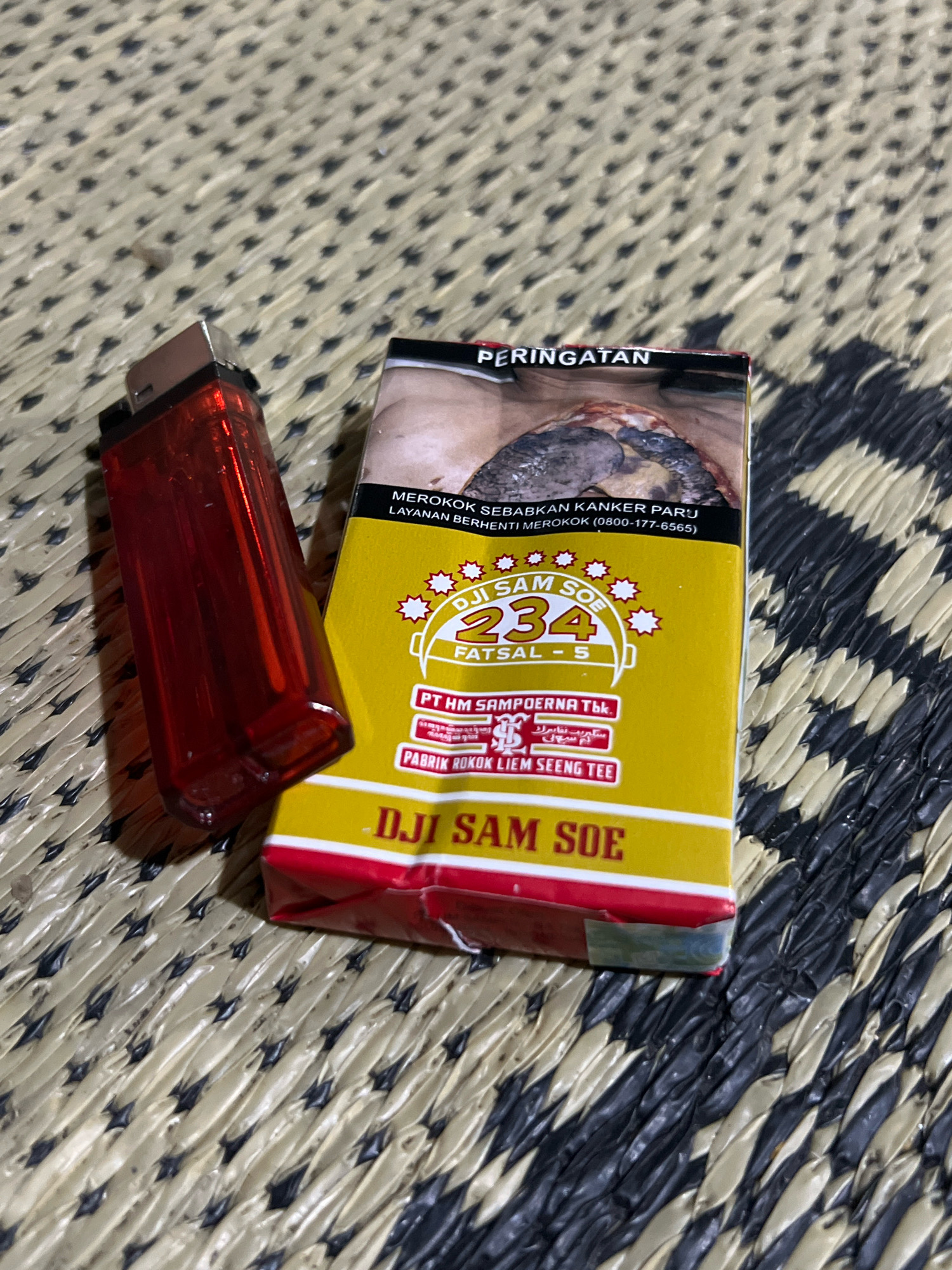 A red plastic lighter is lying next to a yellow cigarette pack on a woven mat. The cigarette pack has the text "DJI SAM SOE 234 FATSAL - 5" and "PT HM SAMPOERNA Tbk". It also displays "PABRIK ROKOK LIEM SEENG TEE". The pack features a health warning in Indonesian that reads "PERINGATAN MEROKOK SEBABKAN KANKER PARU LAYANAN BERHENTI MEROKOK (0800-177-6565)". The image was taken in Tlogoadi, Indonesia.