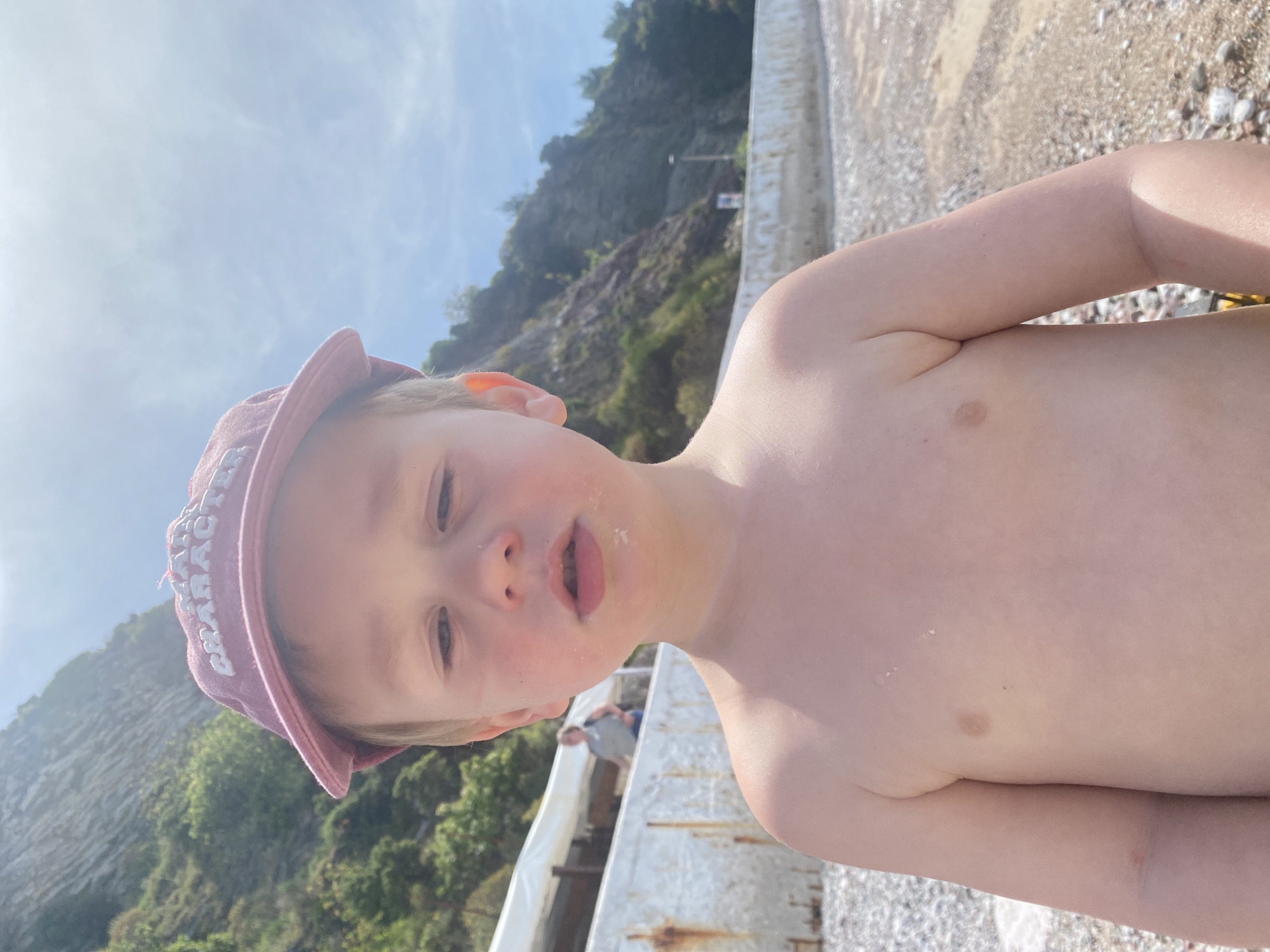 A young, fair-skinned child wearing a red hat with white letters stands shirtless on a pebble beach. The child is facing the camera with sand on their face and body. In the background is a cement structure, and a green, rocky hillside. In the distance, a person is standing under a white canopy. The photo was taken in Rafailovići, Montenegro.
