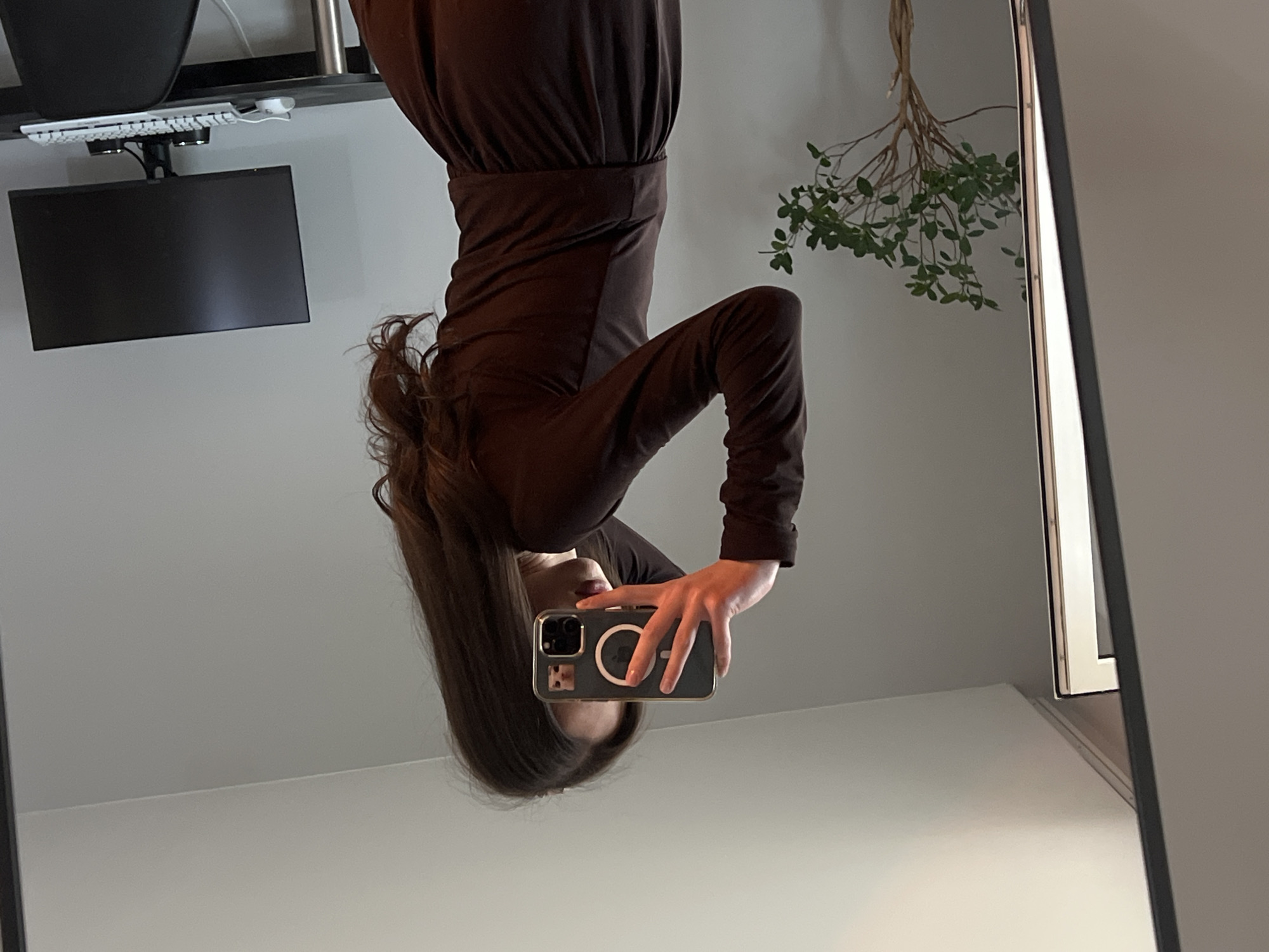 The image displays a mirror selfie taken indoors in Ternopil, Ukraine, presented from an inverted perspective. The main subject is a person with long, dark brown hair, wearing a dark brown, long-sleeved top or dress with a cinched waist. Their face is partially obscured by their hair and the smartphone they are holding up. Light-colored lips are faintly visible.
The person is holding a smartphone with a clear protective case that distinctly shows a white circular MagSafe attachment ring on its back. The hand holding the phone has neatly manicured nails.
The reflected background reveals an indoor setting. To the left (when viewed as the person being upright), there's a dark monitor and a white keyboard, suggesting a desk or workspace area. To the right, a decorative plant with green leaves and thin branches adds a natural touch. The walls are a plain light gray, and a floor or surface line is visible below. The edge of the mirror or reflective panel is seen on the far right side of the frame. The overall aesthetic is clean and modern. There is no discernible text within the image.