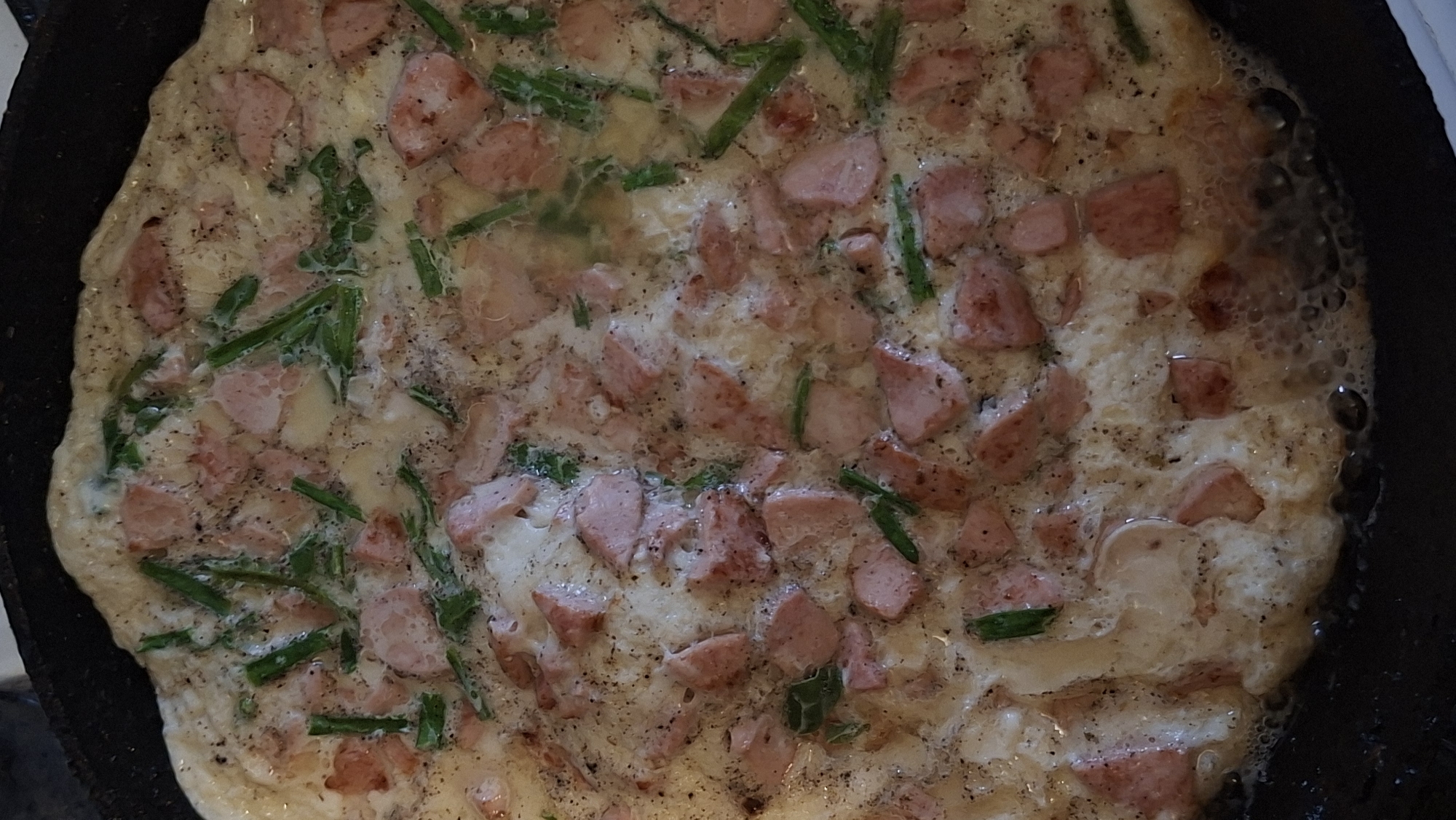 A top-down, close-up shot shows a fully cooked omelet in a black pan. The omelet is a light yellow color and appears to be made with eggs, diced ham, and chopped green onions. The surface of the omelet is uneven and bubbly, indicating it was recently cooked. The pan has a textured surface and a slightly glossy finish. The background is dark and out of focus, suggesting it is a kitchen environment. There are no people or other objects clearly visible in the frame.