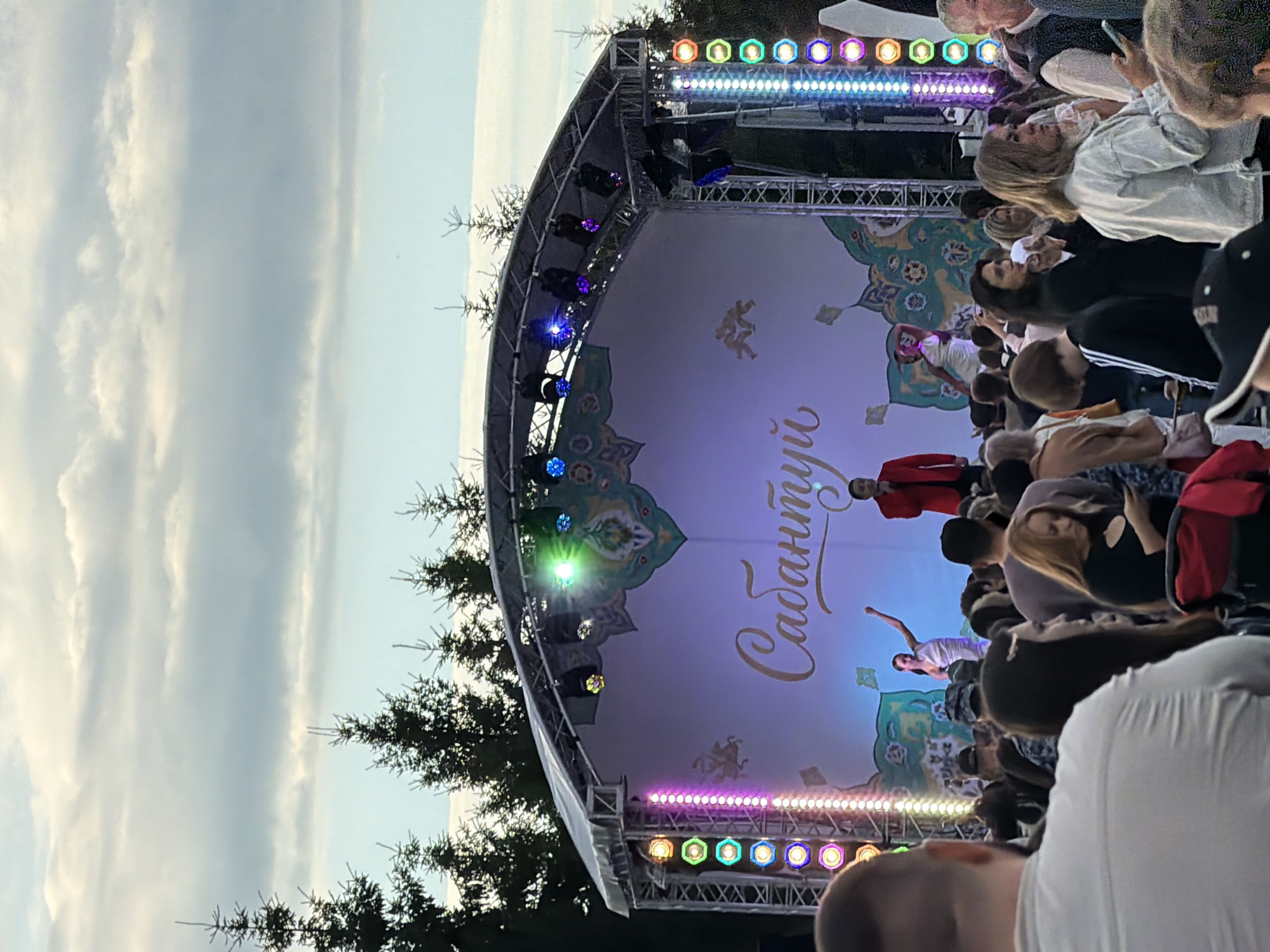 An outdoor stage event is depicted, with a large crowd of people gathered in the foreground and midground. The stage features a white backdrop with colorful decorative elements and Cyrillic text that reads "Сабантуй". There are performers on the stage, including a singer in a red jacket and a dancer in a white dress. The stage is illuminated by colorful lights on both the left and right sides, as well as spotlights on top. Behind the stage, the silhouettes of evergreen trees are visible against a cloudy sky. The audience members are diverse in age and gender, with many looking towards the stage, some taking photos with their phones. The overall atmosphere appears festive.