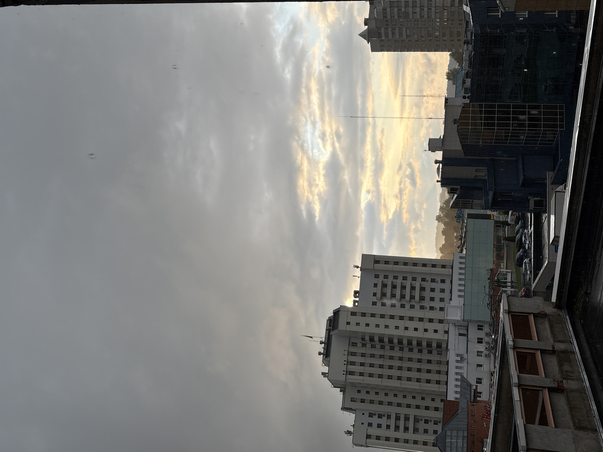 This is an eye-level exterior shot of a city skyline in Vitsebsk, Belarus, under an overcast sky with hints of yellow and orange light breaking through the clouds, suggesting either sunrise or sunset. The foreground shows the tops of buildings with flat roofs, one with exposed concrete framing and another with visible moss. In the mid-ground, a tall, white, multi-story building with many windows dominates the scene, featuring a dark flag atop its roof. To the right of this building, another darker, more modern-looking building with a grid-like facade is visible, and further to the right, a very tall, grey, high-rise apartment building stands. In the background, more buildings and distant hills can be seen. Several cars are parked in a lot in front of a building with a glass facade. There are no people visible in the frame. The overall impression is one of an urban environment with a mix of architectural styles under a dramatic sky.