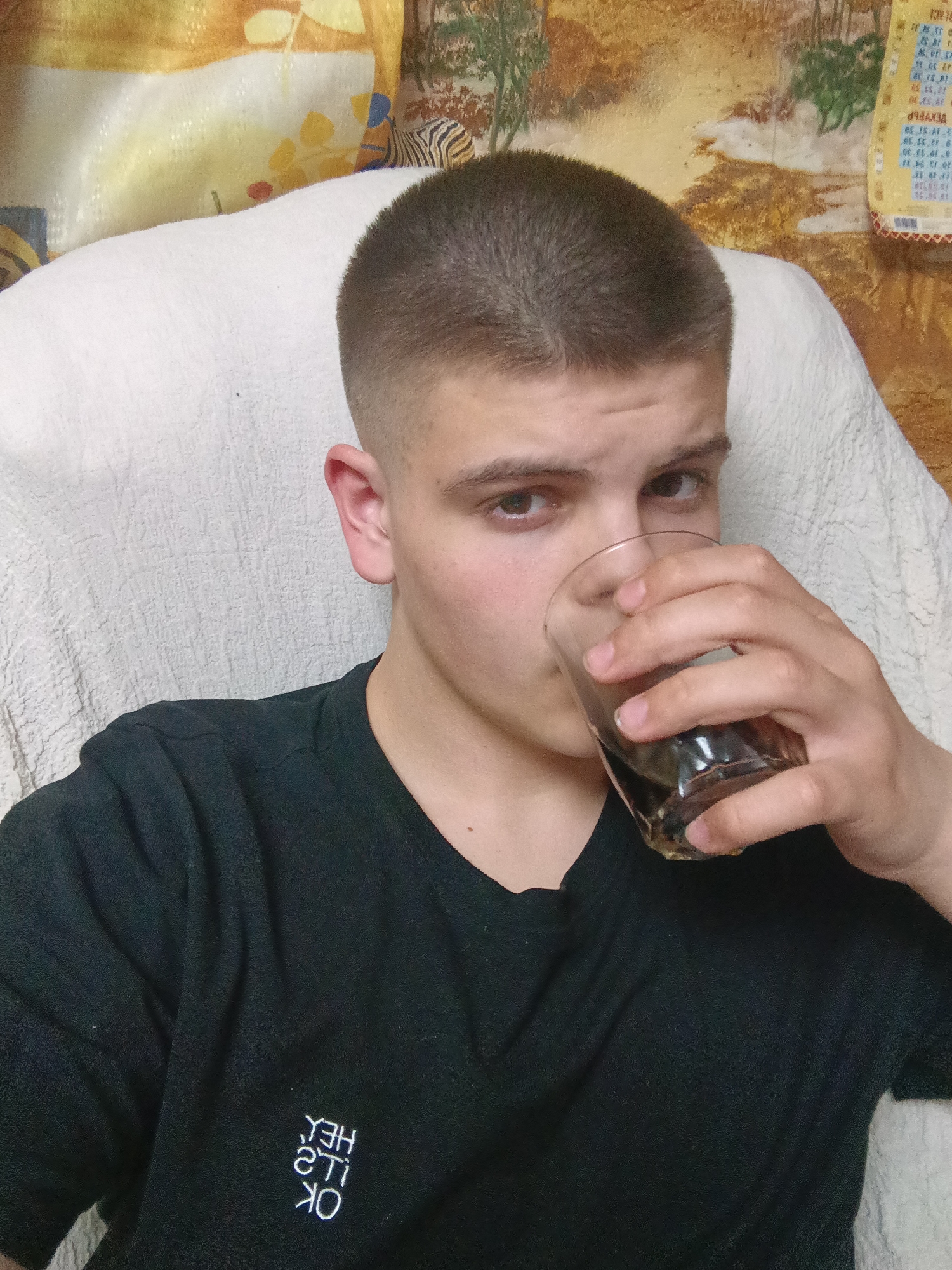 A young man is holding a glass of dark liquid to his lips, seemingly drinking. He has short, cropped hair and is wearing a black t-shirt with white lettering in an upside-down style, located on the chest. The man is seated in a white armchair or large cushion. Behind him is a wall with a patterned wallpaper, including a zebra print and a calendar featuring blue text and numbers on a yellow background. The location is Красный Маяк, Russia. The specific actions occurring are drinking and posing for a picture.