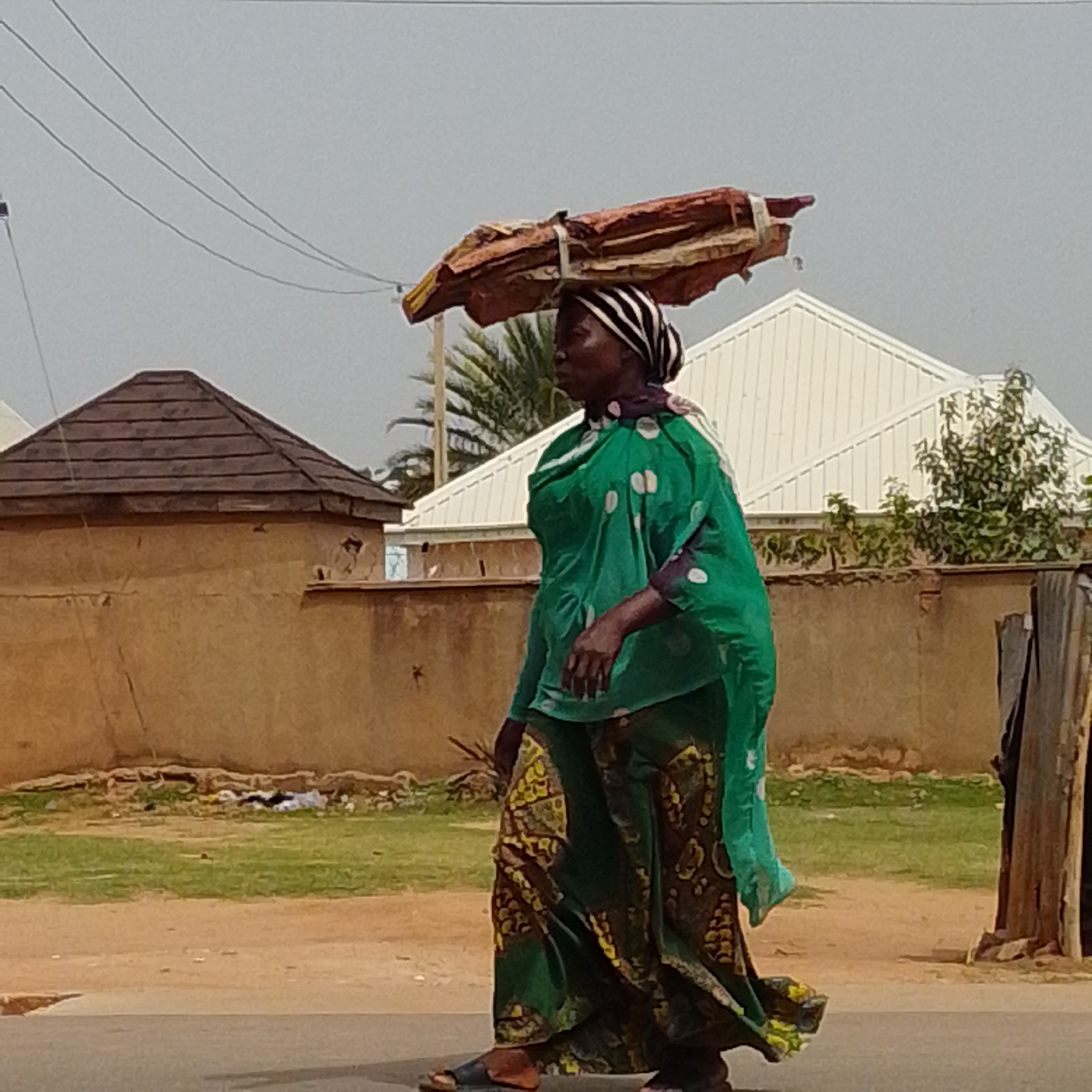 A person walks along a paved street in Gombe, Nigeria, carrying a load of what appears to be dried plant material balanced on their head. The person is wearing a green wrap-around garment with a patterned skirt and a black and white striped headscarf. In the background, there are several buildings with corrugated metal roofs and brown walls. A small patch of green grass with some scattered debris is visible between the street and the buildings. Utility lines are strung across the sky. The overall scene suggests a typical day in an urban environment in Nigeria.