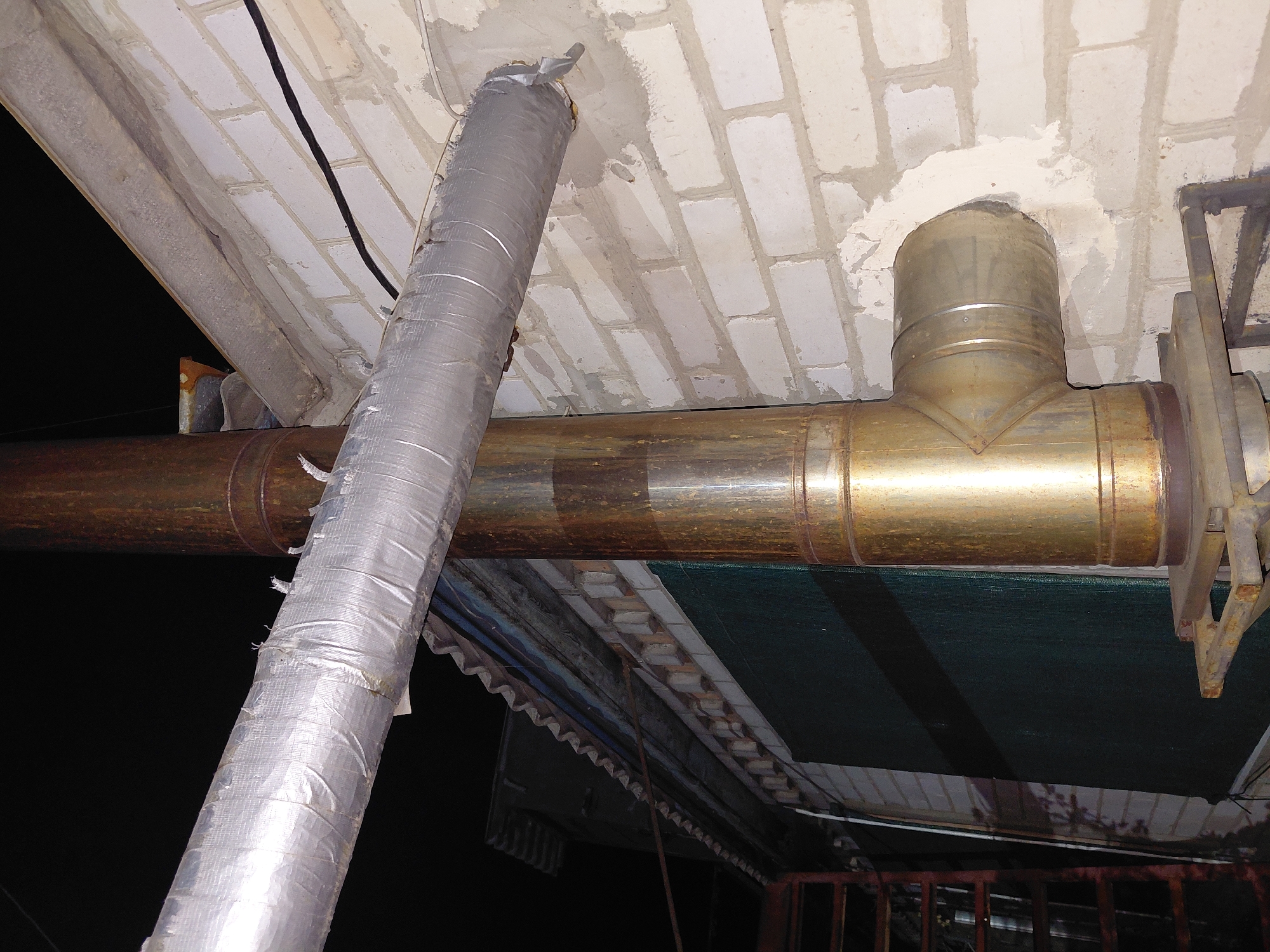 An exterior shot shows a chimney and vent pipe attached to a brick building. The main chimney pipe is vertical and appears to be made of weathered metal, possibly galvanized steel, with a brown patina. A horizontal vent pipe, also metal, extends from the chimney and is wrapped in silver foil insulation tape. The building's wall is constructed of white bricks with visible mortar. A dark-colored cable runs along the top of the brick wall. To the left, a corrugated metal roof structure is visible, and further left, a dark green mesh material is draped over something, with a metal ladder structure partially visible in the foreground. The scene is dimly lit, suggesting it was taken at night or in low light conditions.