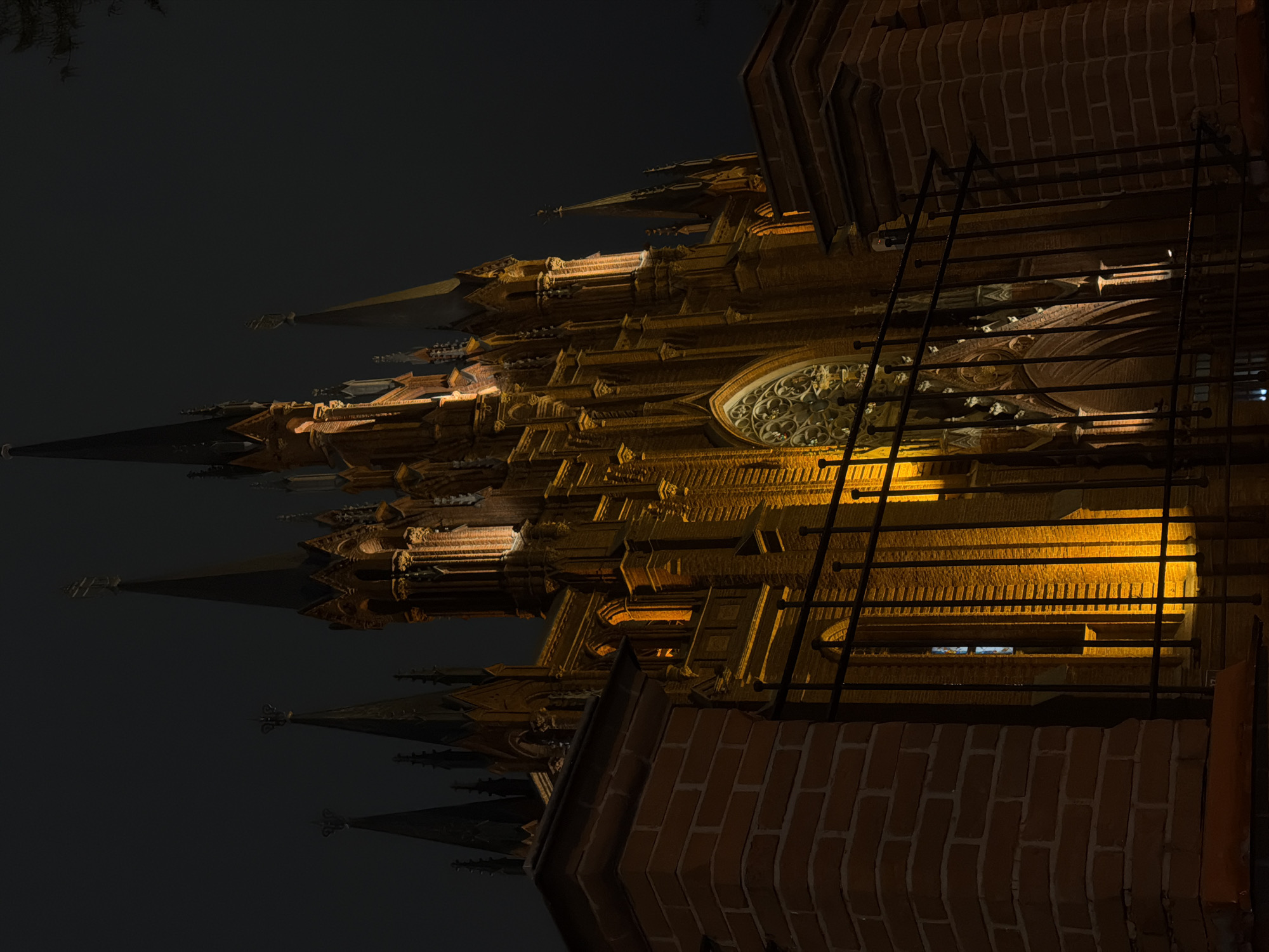 The image shows a building, potentially a cathedral, at night. The structure is ornate with spires and intricate details. It is illuminated with warm, golden light, which contrasts with the dark sky. Part of the building appears to be constructed of brick, and there is a metal structure resembling a ladder or support system attached to it. Based on the architectural style, which features Gothic elements, and the indication of a likely cathedral, the building resembles St. Peter and Paul Cathedral in Moscow, Russia.