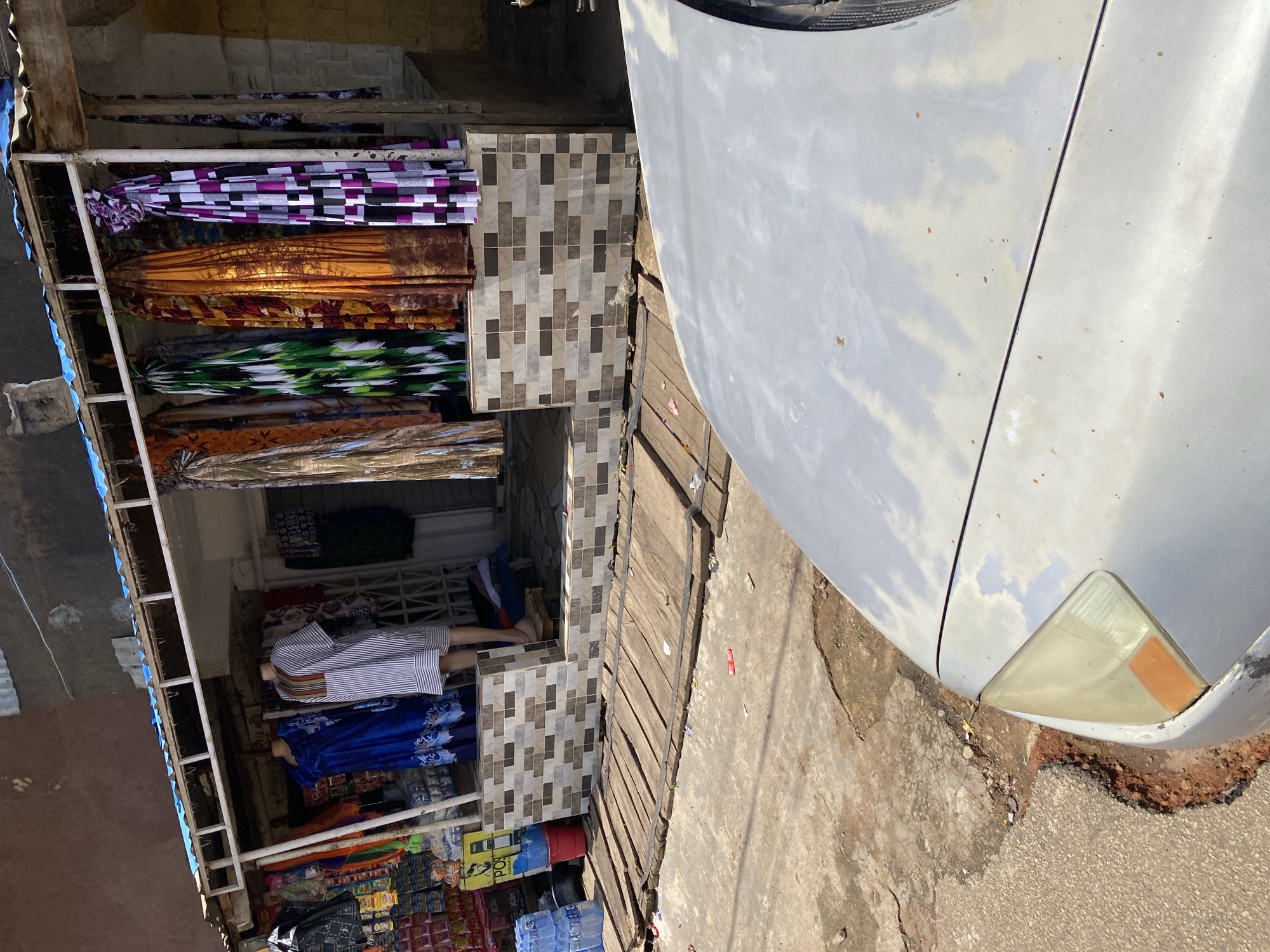 A roadside stall is captured in Jos, Nigeria. The stall features clothing items displayed on mannequins and hung from racks. A blue dress with an intricate pattern is on a mannequin on the left, and a white and brown striped dress is on a mannequin on the right. Several brightly colored fabrics, likely dresses or wrappers, are hung for display. Behind the clothing, shelves are stocked with packaged goods. The stall is set up on a wooden platform, with a tiled base supporting the front of the stall. In the foreground, the hood and headlight of a light-colored car are visible, suggesting the stall is situated near a road. The environment appears to be an outdoor, urban setting with a rough pavement surface and cracked earth visible. The overall impression is a vibrant market scene.