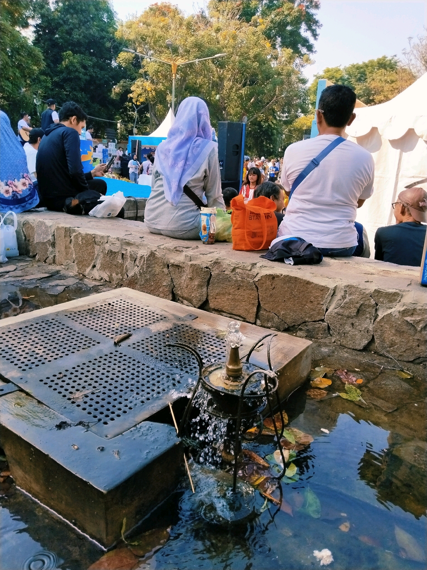 The image depicts an outdoor scene likely within a park or public gathering space in Surabaya, Indonesia. In the foreground, a small decorative fountain with a metallic base is spraying water into a shallow pool. The pool is partially covered by a dark, perforated metal grate. Fallen leaves are scattered in the water and on the surrounding stone ledge.
Behind the fountain, a group of people are seated on a low stone wall, facing away from the camera. One individual in the center is wearing a light purple headscarf and a grey long-sleeved shirt. To their left, a person in a black hoodie is looking at their phone. To the right, a person in a white t-shirt with a blue strap across their chest is seated. Other individuals are visible in the background, some standing and some sitting.
Further in the background, a white tent structure is visible, suggesting an event or market. Blue and white banners are displayed, and there are trees with green foliage. The overall atmosphere appears to be casual and public, possibly during an afternoon gathering or festival. The presence of multiple people and a tent suggests a community event.