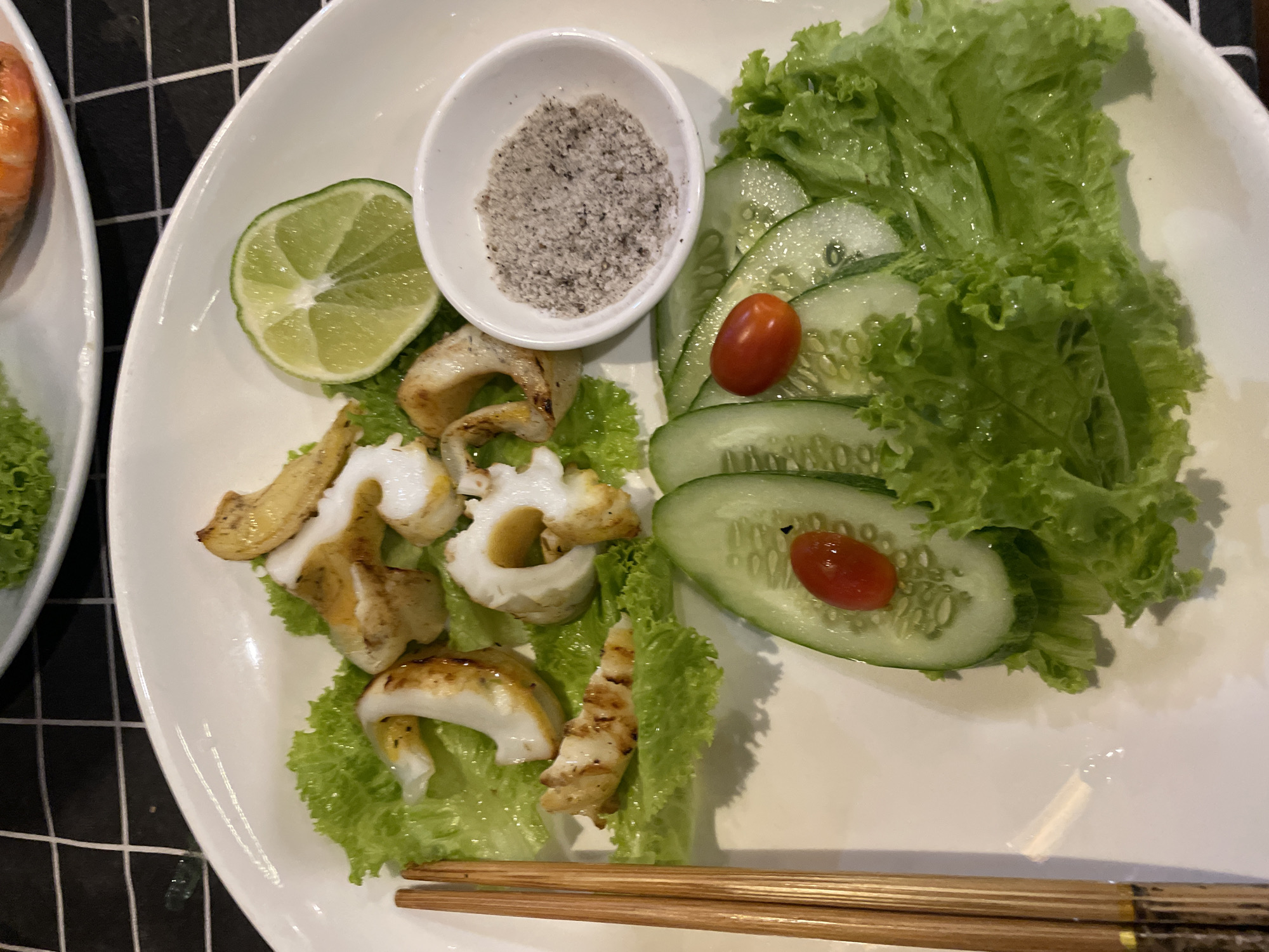 A top-down shot shows a white plate containing grilled squid, lettuce, sliced cucumber, cherry tomatoes, and a lime wedge. A small white bowl of salt is also on the plate. A pair of chopsticks lies at the bottom of the plate. The plate sits on a table covered with a black and white checkered cloth. This scene is likely in a restaurant located in or near Bắc Nha Trang, Vietnam, based on the style of food.
