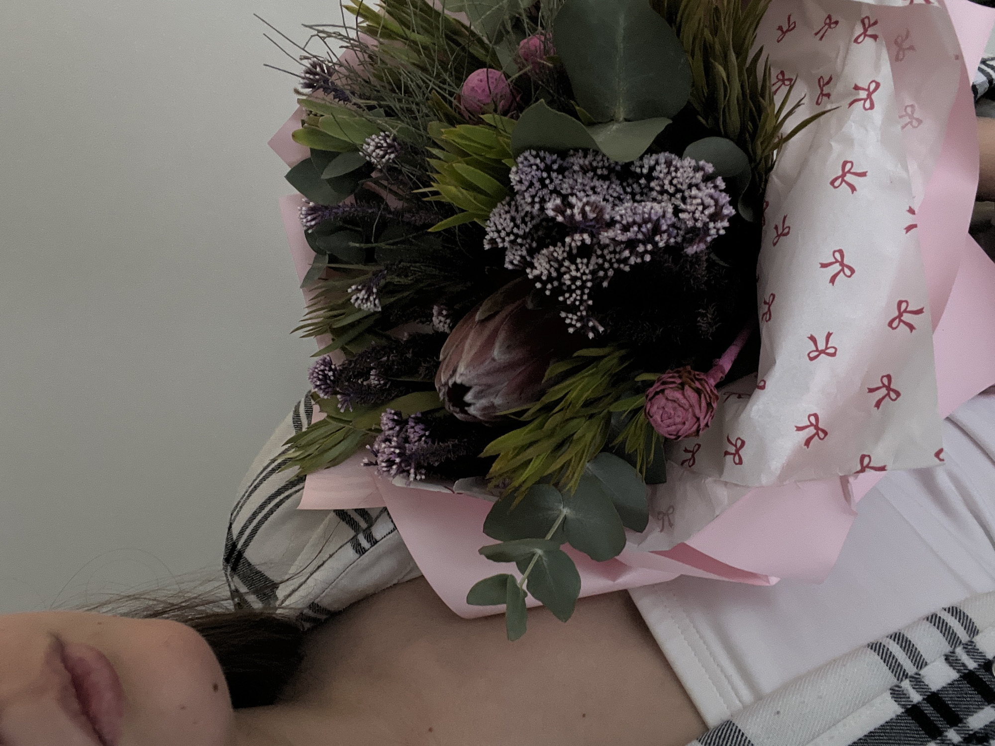The image presents a close-up, angled view of a person alongside a vibrant bouquet of flowers. The person's face, specifically their lips and chin, is visible on the lower left, along with a portion of their dark hair and shoulder. They appear to be wearing a white top with a black and white plaid pattern on the shoulder or sleeve.
The main subject is a lush and textured bouquet, occupying the right and center of the frame. It features a diverse selection of flowers and foliage, showcasing a palette of greens, purples, and pinks. Notable elements within the bouquet include clusters of small, delicate purple-white flowers, a prominent brownish-pink protea-like bloom, smaller spherical pink flowers, and broad, silvery-green eucalyptus leaves. Various other green, spiky, and feathery foliage contribute to the bouquet's rich texture.
The flowers are artfully wrapped in layers of soft pink paper and an inner sheet of white tissue paper, which is decorated with repeating red bow patterns. The background is a plain, light-colored wall, suggesting an indoor setting. The lighting is soft and diffused, creating a serene and intimate atmosphere, possibly indicating a moment of quiet appreciation or reception of the flowers. There is no visible text beyond the decorative bow patterns on the wrapping.