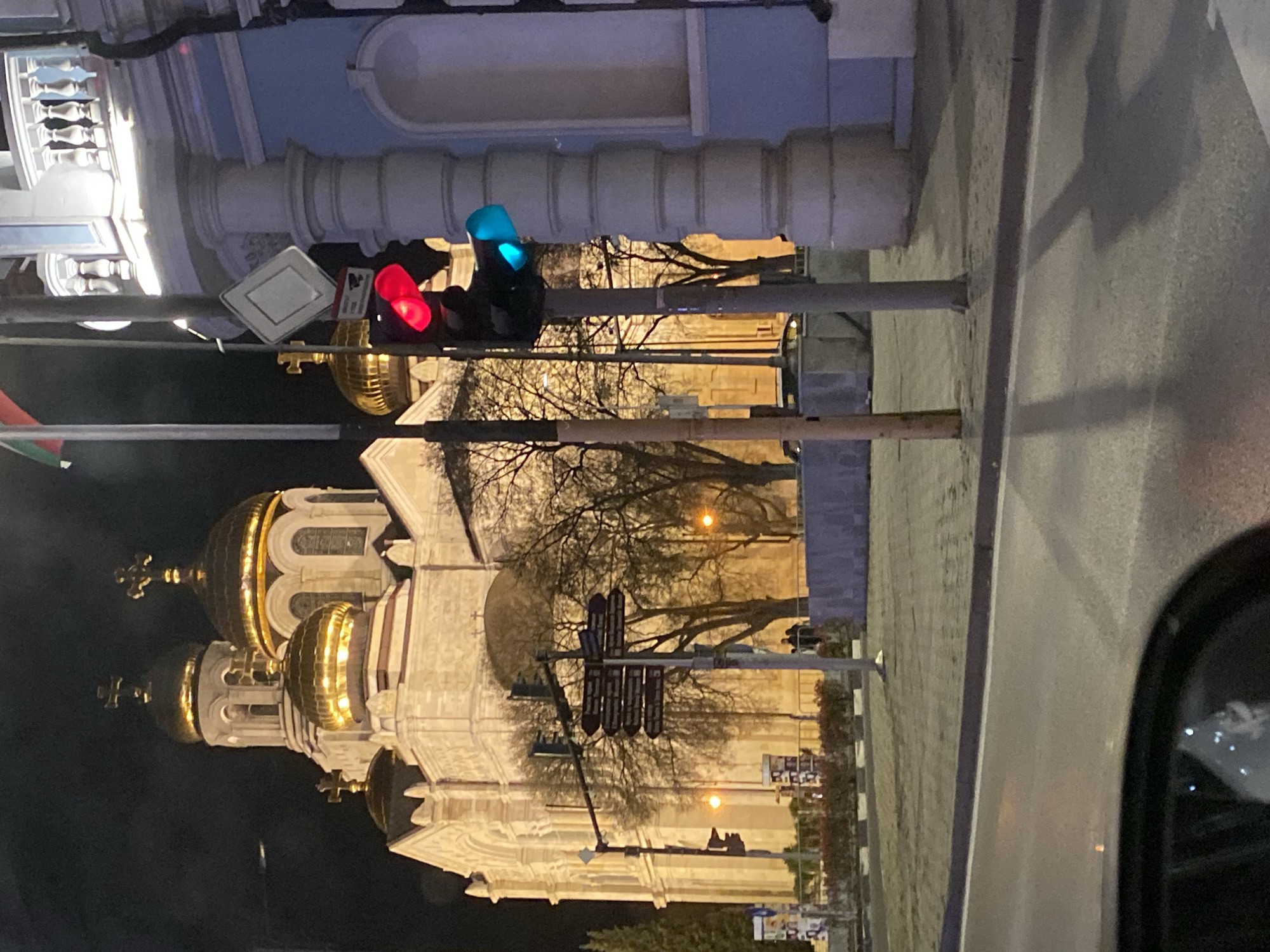 The image shows the Cathedral of the Assumption of the Virgin in Varna, Bulgaria at night. The cathedral's golden domes are illuminated. A traffic light is in the foreground with red and blue lights activated. Bare trees are visible in front of the cathedral. A street is on the right side of the frame, and a car's side mirror is in the lower right corner.