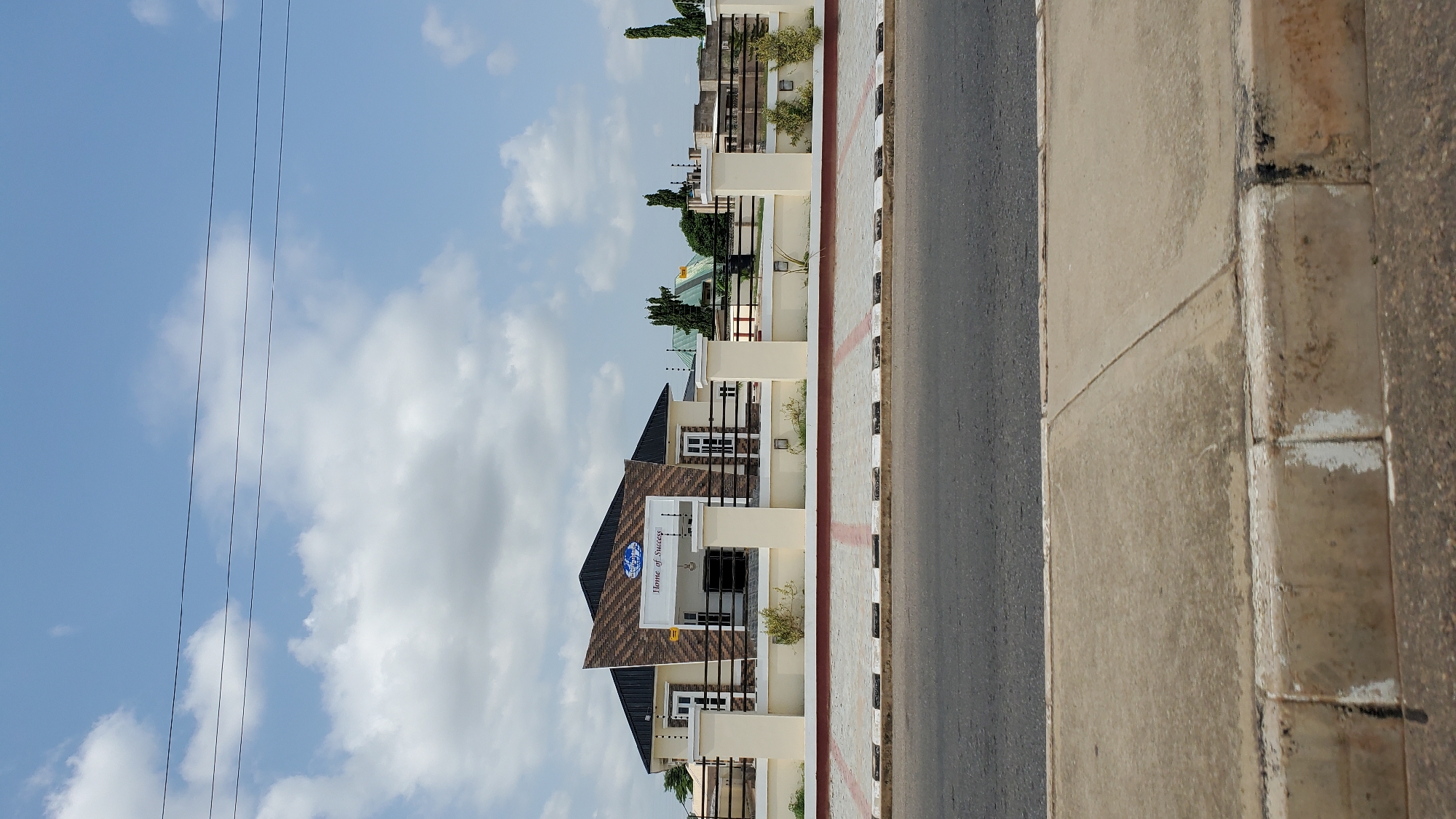 The image shows the "Home of Success" building, a modern, single-story structure with a dark roof and a distinctive brown and beige patterned facade above the entrance. The building is enclosed by a light beige wall with decorative pillars and a dark metal fence. Small green plants are visible along the base of the wall inside the fence. In front of the building is a paved walkway with a black and white checkered border. A paved road runs in front of the walkway. The sky above is blue with scattered white clouds. The scene is consistent with an urban environment in Jalingo, Nigeria.