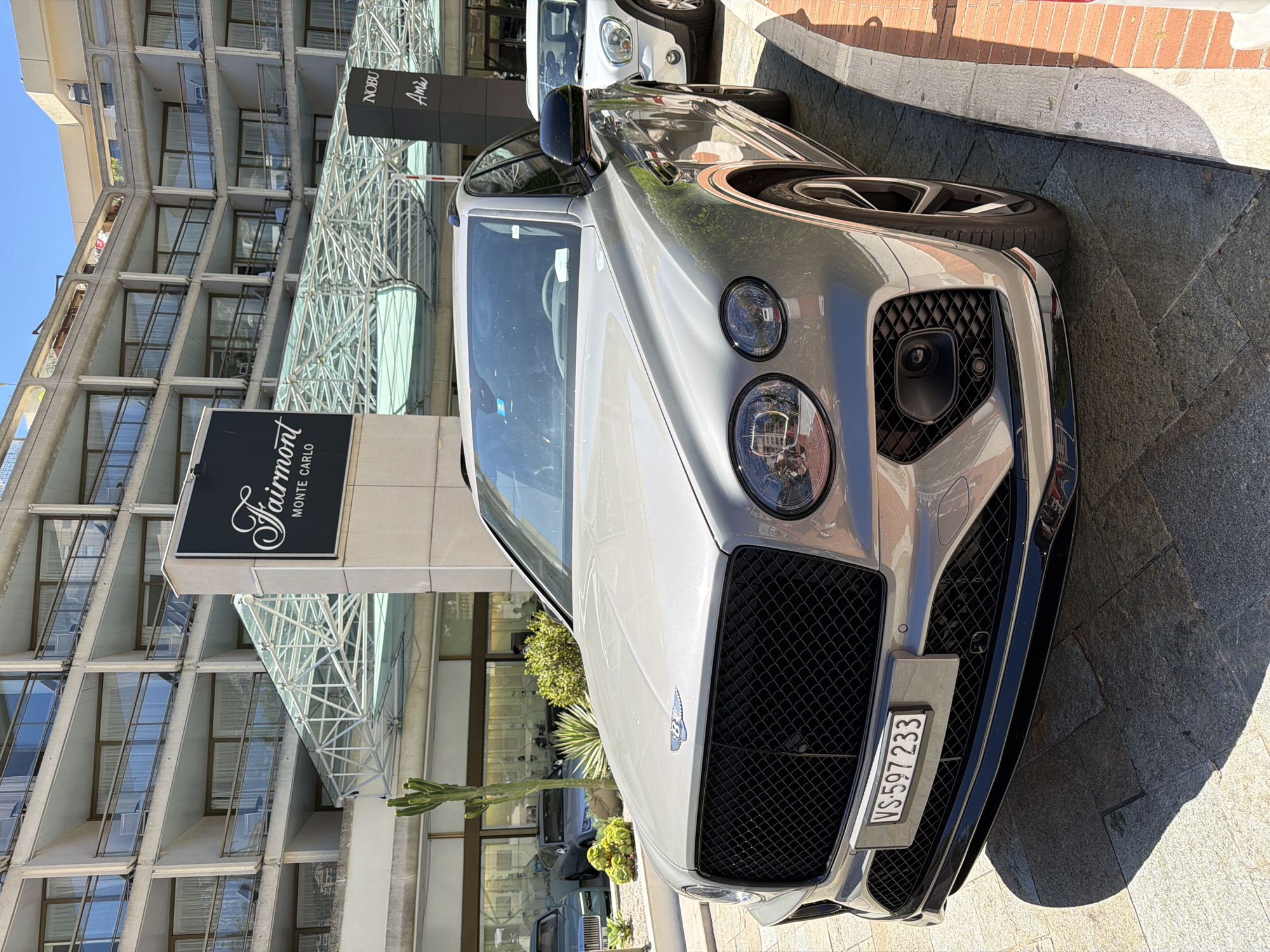 The image captures a silver Bentley Bentayga parked on a paved street in front of the Fairmont Monte Carlo hotel in Monaco. The hotel, a prominent landmark in the city, features a distinctive brutalist architectural style with multiple levels of balconies and glass facades. Adjacent to the Fairmont signage, a "NOBU Ami" sign is visible, indicating a restaurant or establishment within the hotel complex. Several other vehicles are present, including a white car behind the Bentley and other cars parked in the background. A cactus and various plants are arranged in planters near the hotel entrance. The scene is bathed in sunlight, casting shadows on the ground. The license plate of the Bentley reads "VS 597 233". There are no visible people in the immediate foreground.