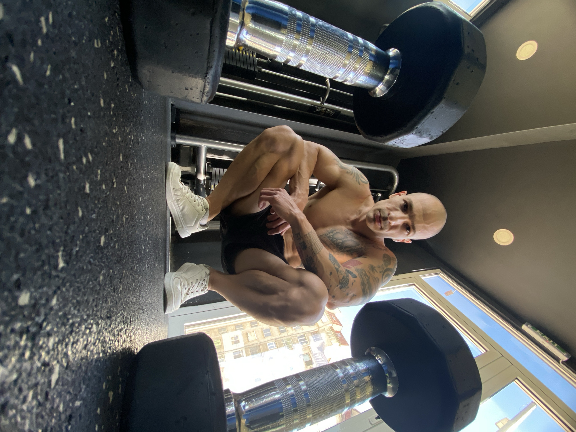 A bald man with tattoos is crouching on the floor of a gym between two dumbbells. He is wearing white sneakers and black shorts. The floor is speckled and the ceiling is dark. Through a window, a building is visible, possibly located in Pleven, Bulgaria.