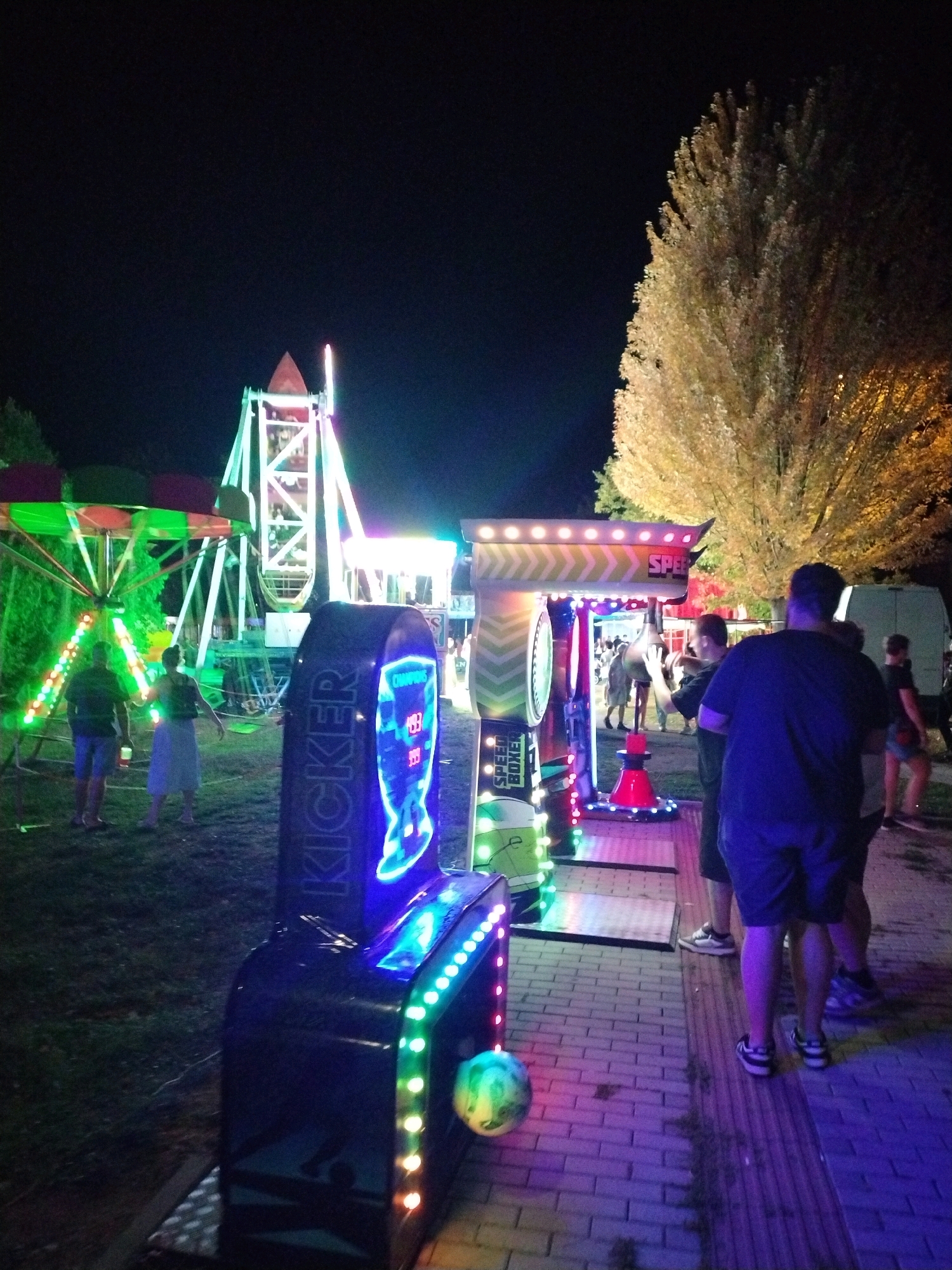 At night, on a paved area with grass to the left, an amusement area is depicted. In the foreground, a brightly lit arcade game, labeled "KICKER" in neon blue letters, is partially visible. To the right of the Kicker game, another arcade game, the "SPEED BOXER", is active with a person striking it. Further in the background, a Ferris wheel with multi-colored lights and a smaller amusement ride with green and pink accents are set up. Several people are scattered throughout the scene, some observing the games, while others are walking. A large, illuminated tree with yellow-gold foliage stands prominently on the right side of the image. In the far background, a white van is parked. The overall atmosphere is festive and illuminated by the various lights of the amusement rides and games. The scene appears to be part of a temporary fair or carnival event, possibly in Písek, Czechia, given the context.