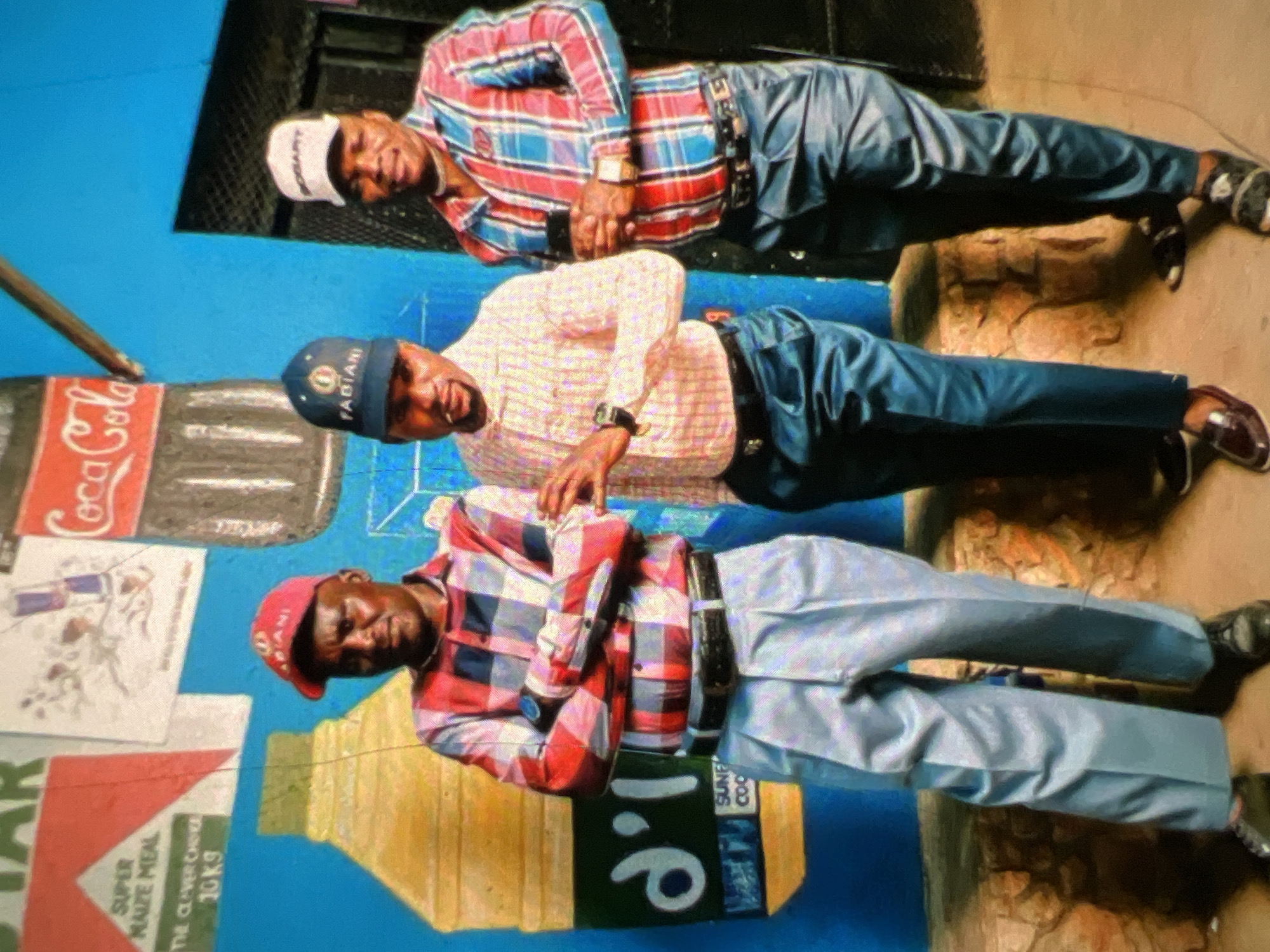 Three men stand in front of a vibrantly painted blue wall with various advertisements. The man on the left, wearing a red, white, and blue plaid shirt, light blue trousers, and a red baseball cap, has his arms crossed. He has dark skin and appears to be middle-aged. To his right, a younger man with dark skin wears a light yellow and white striped shirt, dark blue satin trousers, and a blue baseball cap. He is holding a cigarette and has his right hand raised. The man on the far right, also with dark skin, wears a red, blue, and white plaid shirt, dark blue trousers, and a white baseball cap. He holds a dark rectangular object in his right hand and has a watch on his left wrist. The wall behind them features advertisements for "SUPER MAIZE MEAL" and a Coca-Cola bottle. The ground appears to be dirt and rubble, suggesting an outdoor, possibly informal commercial setting in Lagos, Nigeria.