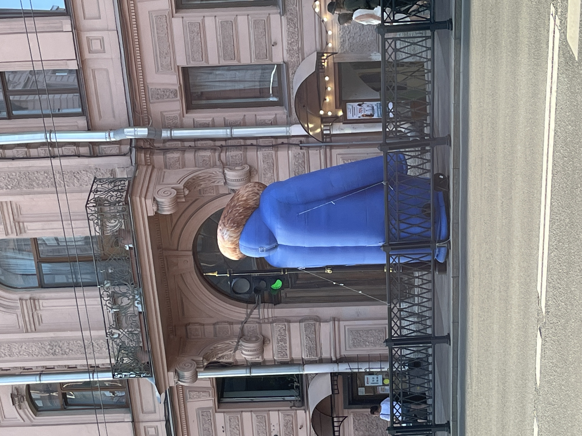 An inflatable sculpture resembling a person in a blue jacket with a brown hood is positioned on a sidewalk in front of a building in Saint Petersburg, Russia. The building, with its pink facade and ornate architectural details, appears to be an apartment building or a commercial establishment. A traffic light with a green signal is visible to the left of the sculpture, near an entrance. A black metal fence separates the sidewalk from the street. Several individuals are present on the sidewalk, some walking and others standing. The overall scene suggests an urban environment on a bright, possibly sunny day.