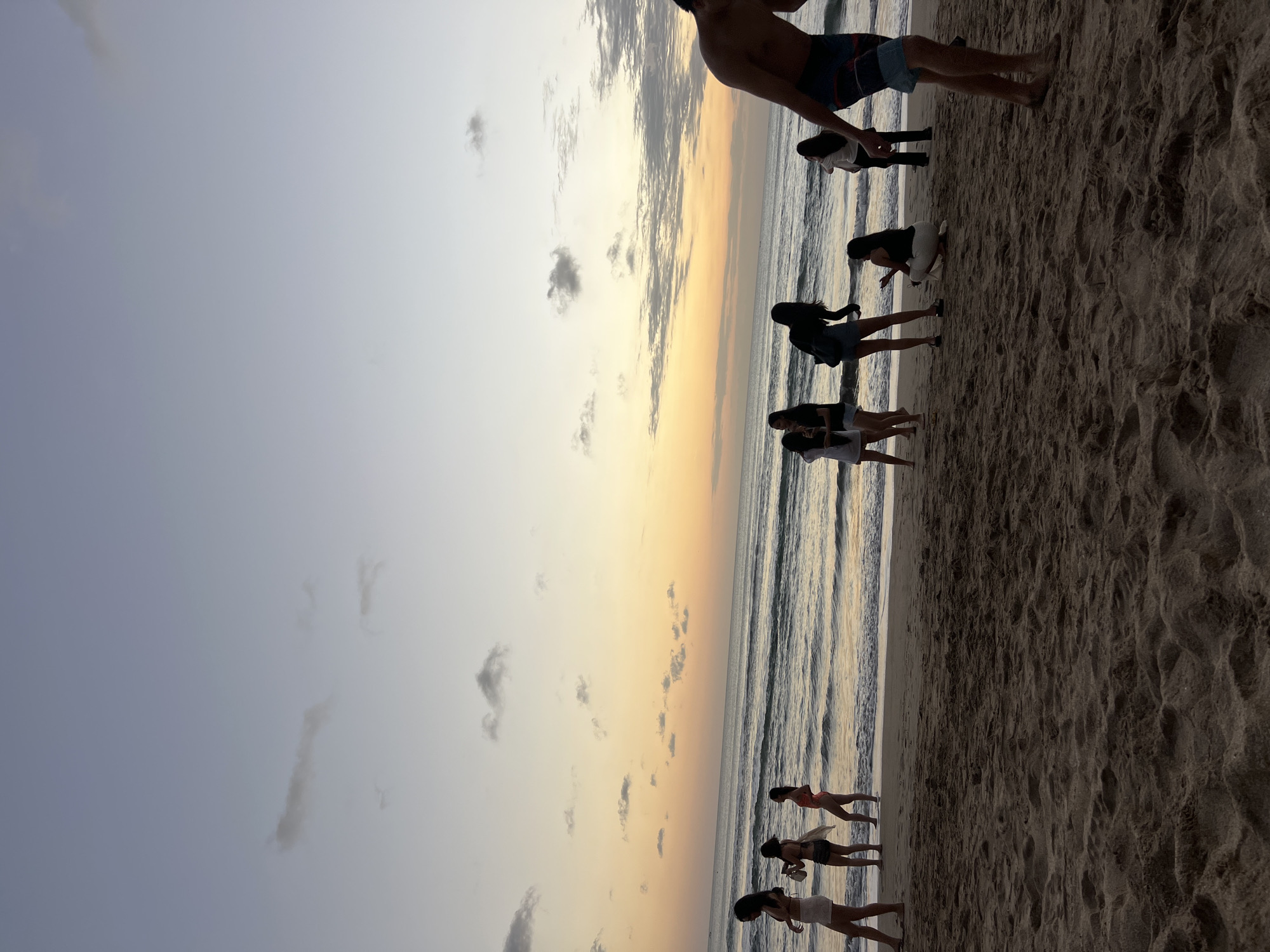 This is a low-angle, medium shot of a beach in Kuta, Indonesia, during what appears to be sunset or dawn. The sky is a gradient of pale blue and yellow-orange, with scattered clouds. The ocean is calm, with gentle waves breaking on the shore. A group of people are scattered across the sandy beach, some standing by the water's edge and others further back.
In the foreground, the sand is textured with footprints and small indentations. To the right, a shirtless man wearing blue and white patterned swim trunks is visible from the waist down, walking away from the camera.
Further back, near the water, several individuals are present. From left to right, a woman in a light-colored top and white shorts stands with her back mostly to the camera. Next to her, another woman in a bikini top and denim shorts holds a light-colored object. A third woman, further to the right, wears a pink bikini.
In the middle ground, several more people are visible. A woman in a dark top and shorts stands with her back to the camera, facing the water. To her right, a woman in a white top and dark shorts is slightly turned. Another woman is crouched down on the sand, wearing a dark tank top and white pants.
Towards the right side of the middle ground, a group of three individuals stand near the water, their backs to the camera. One is wearing a white top, and the other two are in dark clothing.
The overall scene depicts a casual gathering of people enjoying the beach environment.