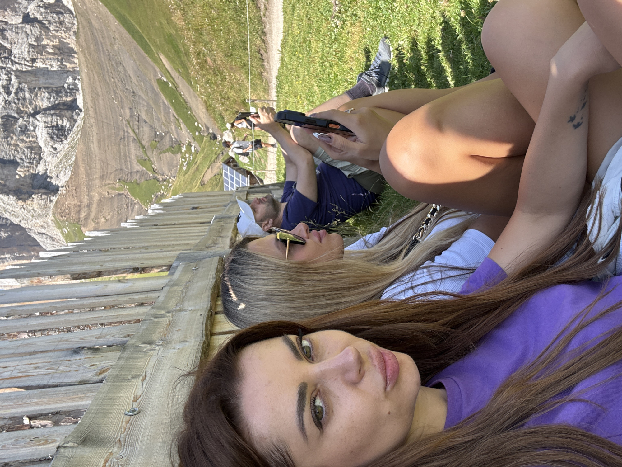 Three people are sitting on the grass in front of a wooden fence, with a mountain landscape in the background. The person in the foreground, on the left, is a woman with long brown hair, wearing a purple t-shirt. She is looking at the camera. To her right is another woman with blonde hair, wearing sunglasses and a white top. She is looking to the right. Behind them, a man wearing a white baseball cap and a dark blue t-shirt is sitting on the grass, holding a smartphone. He appears to be looking at his phone or a screen. In the background, two other figures, possibly hikers, are visible further up a grassy slope with a rocky mountain face behind them. There are some solar panels visible to the left of the man. The location is identified as Campitello di Fassa, Italy, suggesting a mountainous region. The weather appears to be sunny.