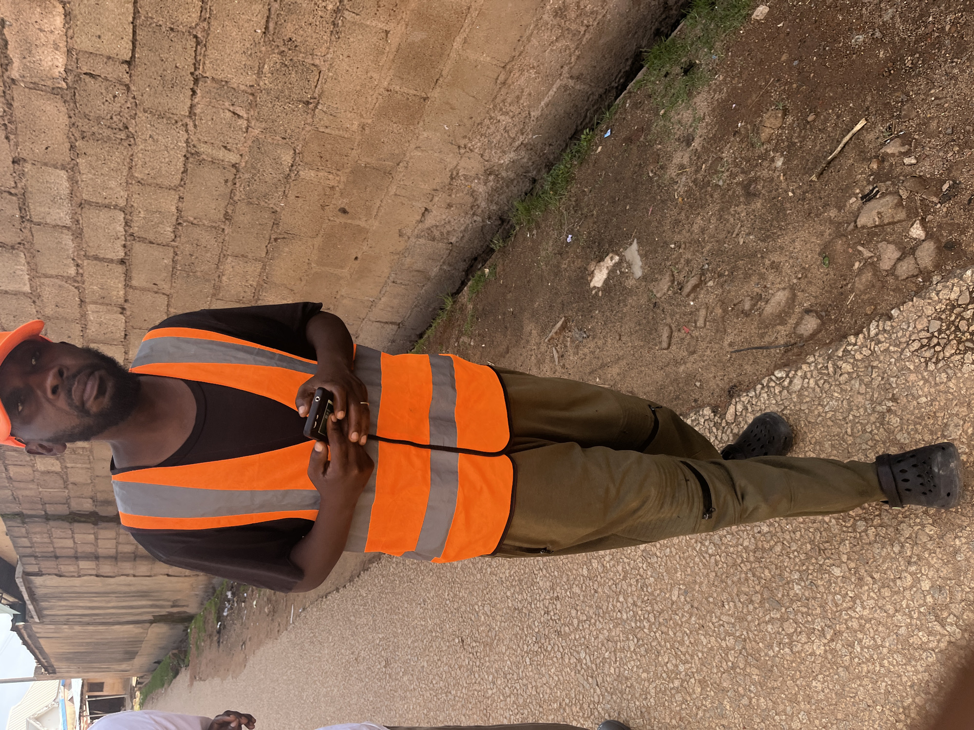 In the city of Jos, Nigeria, a man wearing an orange reflective vest over a black t-shirt and green cargo pants stands on a paved path beside a brick wall. He is holding a mobile phone in his hands. His footwear consists of black crocs. The environment appears to be an outdoor urban setting with a narrow drainage ditch running along the base of the brick wall. The ground is a mix of dirt and some gravelly paving. A second person, partially visible in white clothing, is to the left of the frame. The lighting suggests it is daytime.