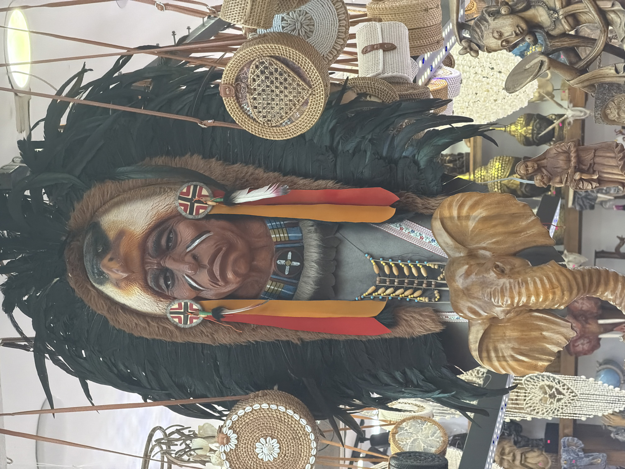 This media file captures a close-up view of an eclectic display of decorative items, likely in a shop or market stall in Fethiye, Turkey. The central and most prominent subject is a large, ornate mask or headdress resembling a Native American chief. It features a brown, carved face with painted white markings, a faux fur trim, and a substantial black feather headdress extending outwards. Two circular medallions with a red cross on a white background, bisected by black lines, are attached near the face, from which hang strips of red and yellow fabric and a single white feather with black tips.
Surrounding this central piece, and extending into the background, is a dense arrangement of various handicrafts. Numerous woven baskets and bags of different sizes and styles are visible, some round with intricate patterns or shell decorations, and others more cylindrical. To the right of the headdress, a large, carved wooden elephant head with detailed ears and a trunk is displayed. Further back, several other wooden carvings can be seen, including a figure resembling a woman holding a child, and an ornate, human-like sculpture. Glimpses of golden Buddha statues are also present in the background.
The setting appears to be indoors, indicated by the artificial light source visible in the top left corner and the tightly packed arrangement of goods. Leather straps are strung across the top of the frame, possibly used for hanging some of the items. No specific activity is taking place; the image depicts a static display of merchandise. The collection of items, ranging from a Native American-style headdress to an elephant carving and Buddha statues, suggests a diverse inventory aimed at tourists or a broad clientele. There is no visible text in the image.